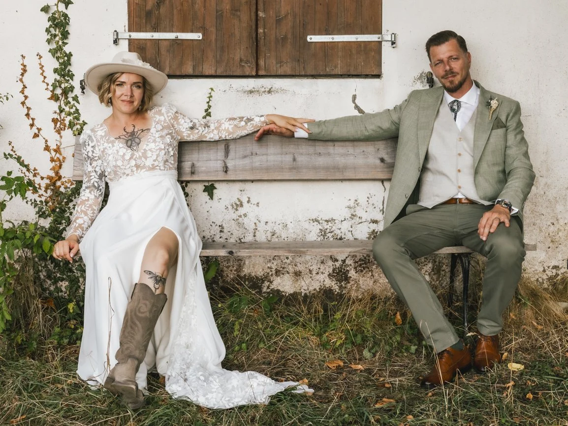Portrait d'un couple de mariés au style bohème assis sur un banc rustique devant une grange, se tenant la main, réalisé en lumière naturelle lors d'un mariage en Suisse