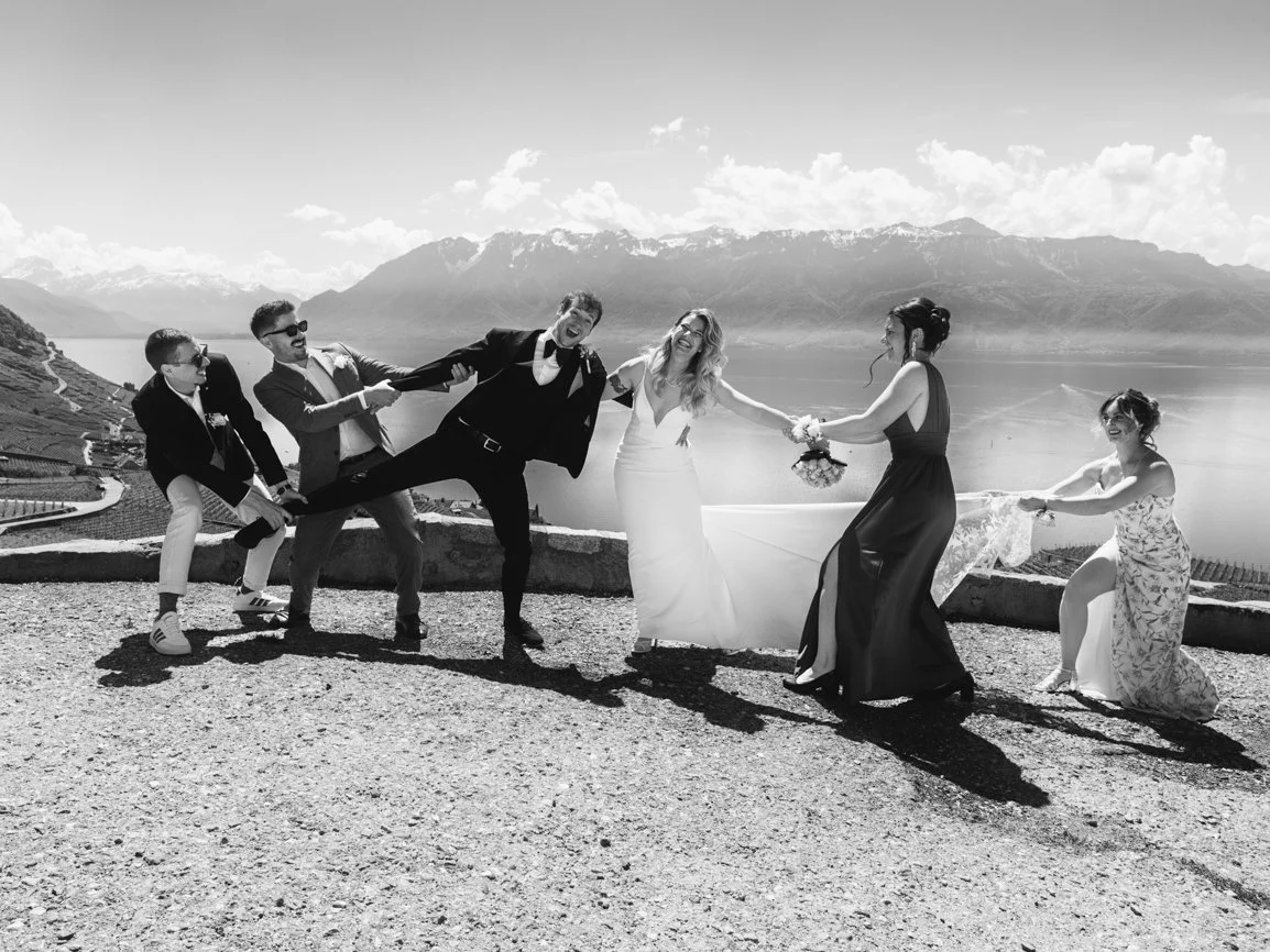 Photo de groupe de mariage originale et pleine d'humour où les mariés et leurs proches jouent devant le lac Léman et les Alpes, capturant un moment spontané et mémorable en Suisse.