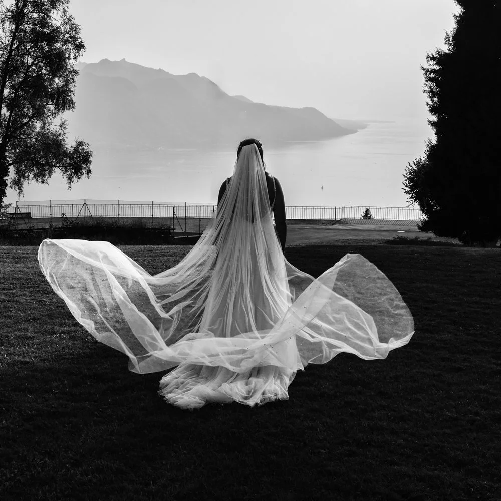 La mariée de dos face au lac Léman et aux Alpes, son long voile porté par le vent, dans une photographie de mariage artistique réalisée à Lavaux en Suisse.