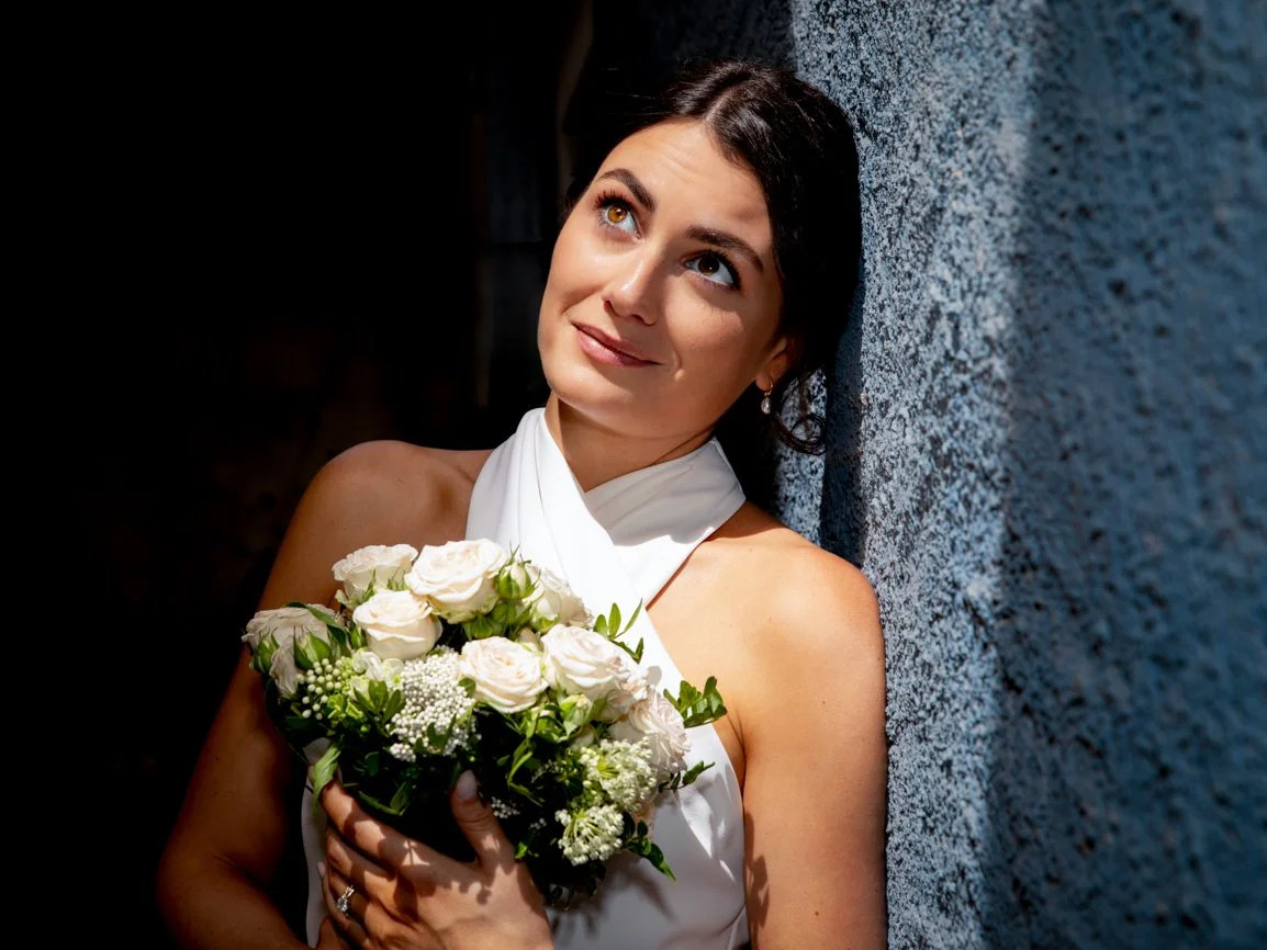Portrait lumineux d'une mariée tenant son bouquet de roses blanches, appuyée contre un mur en pierre, réalisé en lumière naturelle lors d'un mariage en Suisse