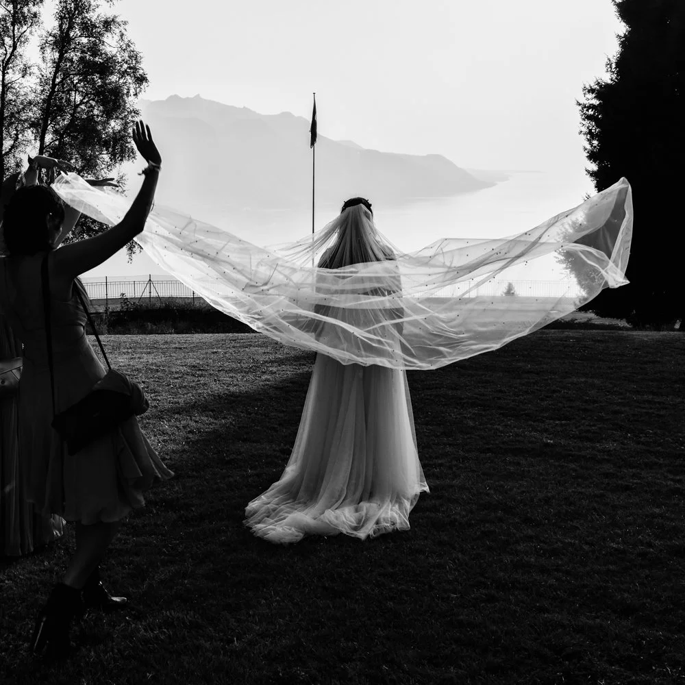 La mariée de dos avec son voile déployé face au lac Léman et aux Alpes, dans une photographie de mariage en noir et blanc réalisée à Lavaux en Suisse.