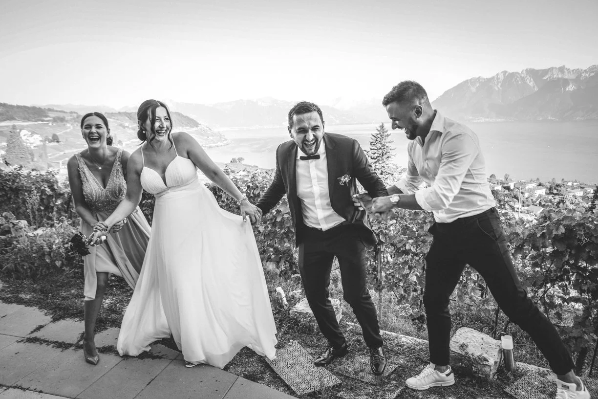 Les mariés éclatent de rire avec leurs témoins au cœur des vignobles de Lavaux, photographie de mariage en noir et blanc capturant une scène spontanée, pleine de joie et de complicité avec le lac Léman et les Alpes en arrière-plan
