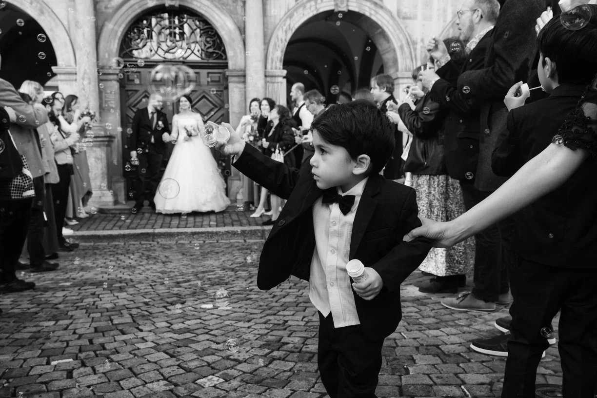 Jeune garçon soufflant des bulles de savon lors de la sortie des mariés devant l'église, photographie de mariage en noir et blanc capturant un instant spontané, vivant et rempli d'émotion en Suisse.