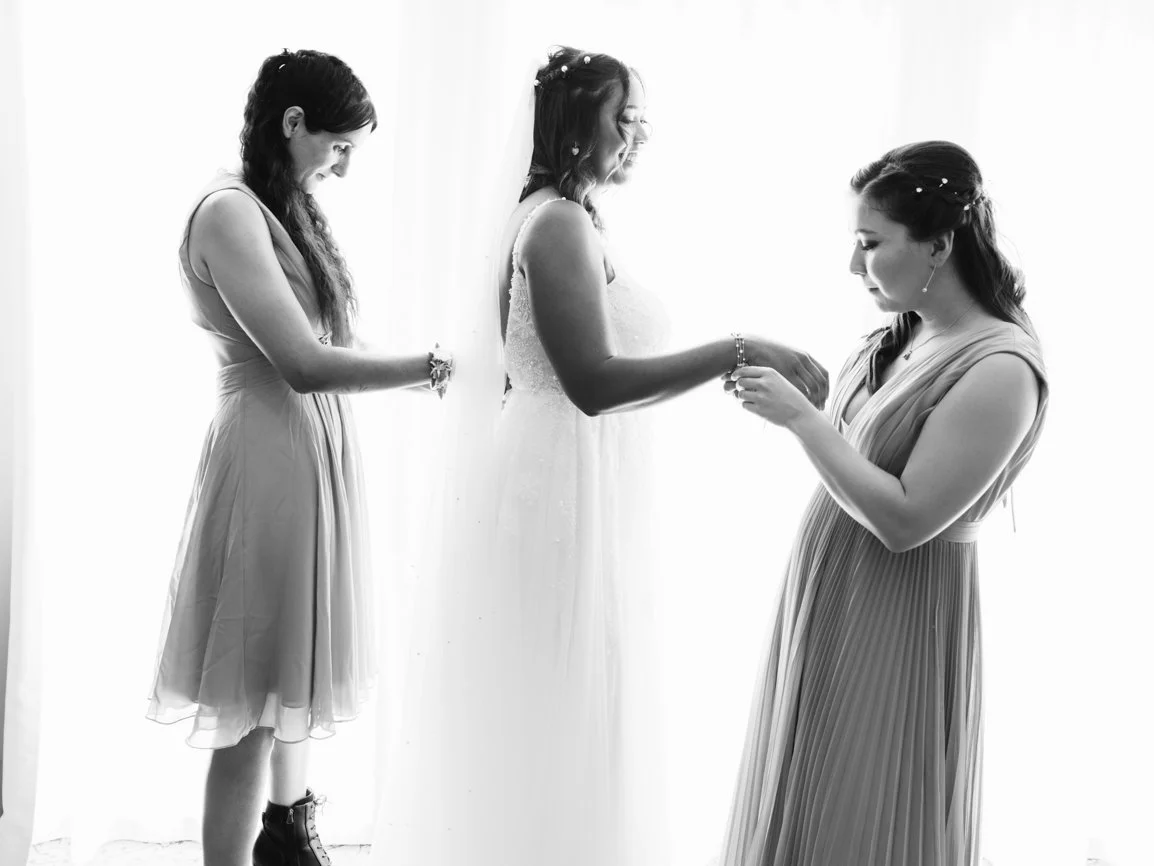 Les demoiselles d'honneur aident la mariée à enfiler son bracelet pendant les préparatifs du mariage, dans une photo en noir et blanc lumineuse mettant en valeur un moment de complicité.