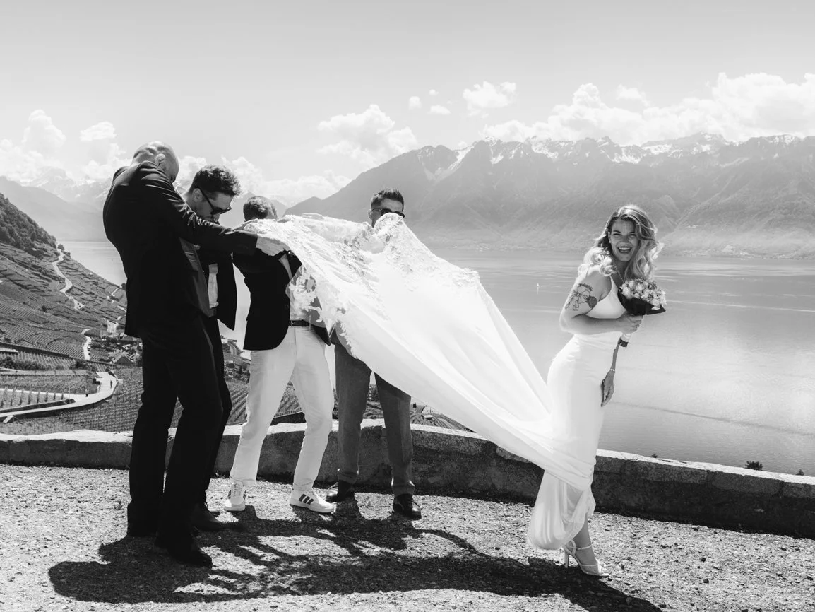 Photo spontanée et pleine d'humour où le voile de la mariée est emporté par le vent tandis que les invités tentent de le retenir, avec les Alpes et le lac Léman en arrière-plan lors d'un mariage en Suisse