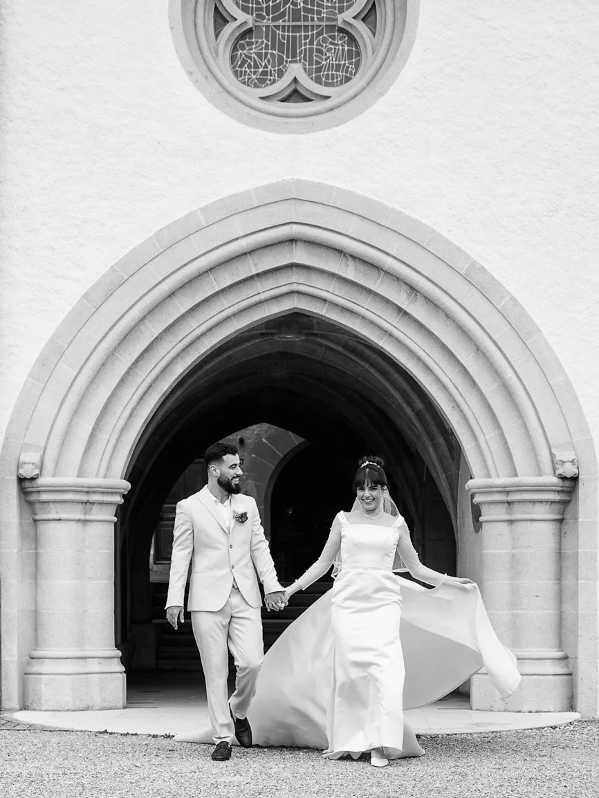 Les mariés marchent main dans la main sous les arches d'un château, la robe de la mariée en mouvement, photographie de mariage en noir et blanc capturant un instant élégant et spontané en Suisse.