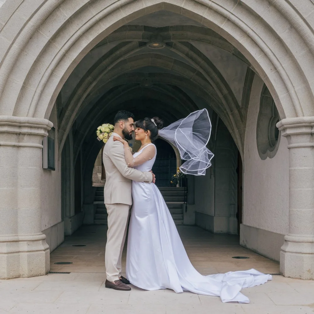 Couple de mariés enlacés sous une arche en pierre avec le voile de la mariée porté par le vent, photographié en Suisse.