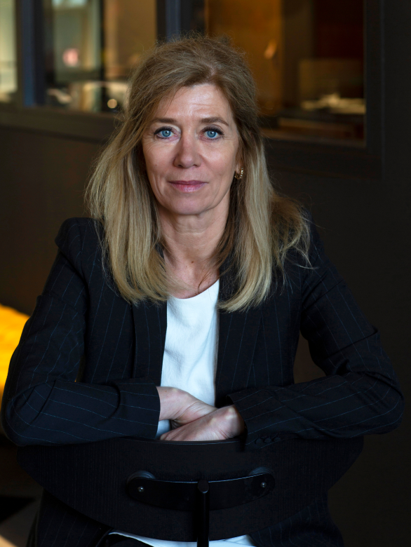 Fotografie van een vrouw met blond haar en blauwe ogen, zittend op een stoel, gekleed in een zwart gestreept jasje en witte blouse, binnen in een kantoor of openbare ruimte.
