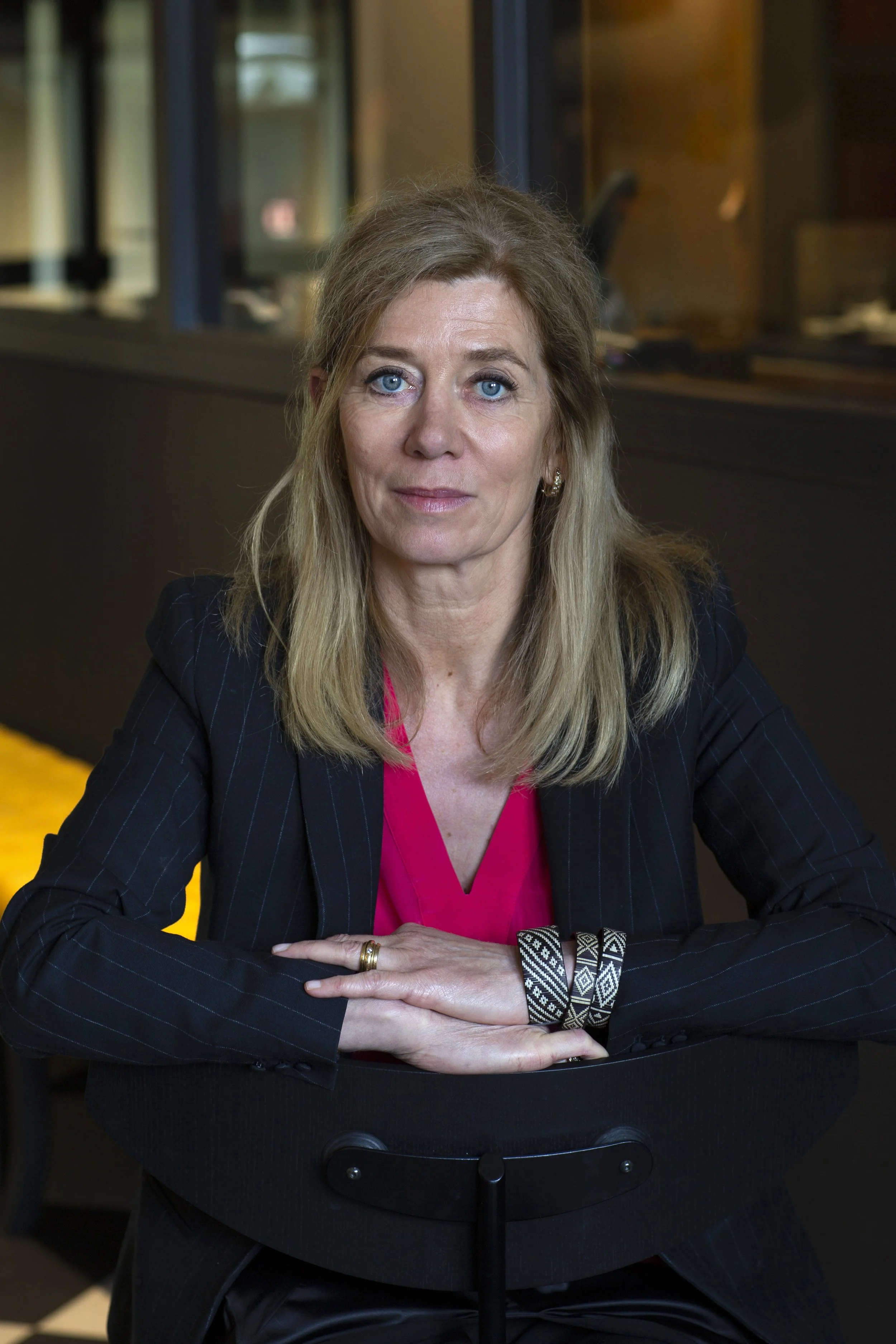 Een vrouw met lang blond haar, blauwe ogen en een nagellak met zwarte en witte patronen, zittend met haar armen gevouwen op een tafel.