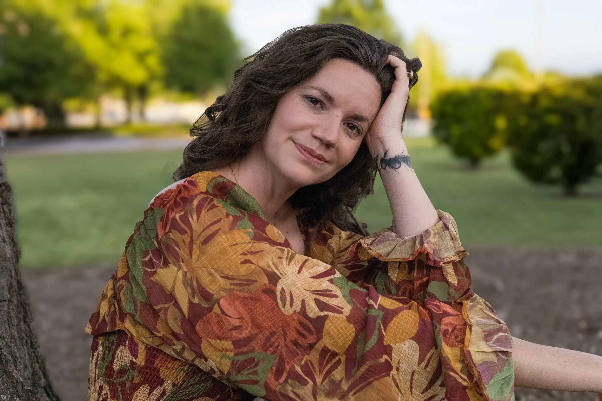 A woman with dark wavy hair rests her head on her hand, sitting outdoors in a park with green trees and blurred background.