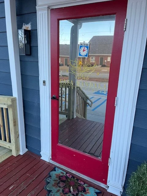 Red door with a glass panel on a blue building, with a decorative butterfly and the text 'Casa Mandala Holistic Center' on the glass, and a floral doormat in front.
