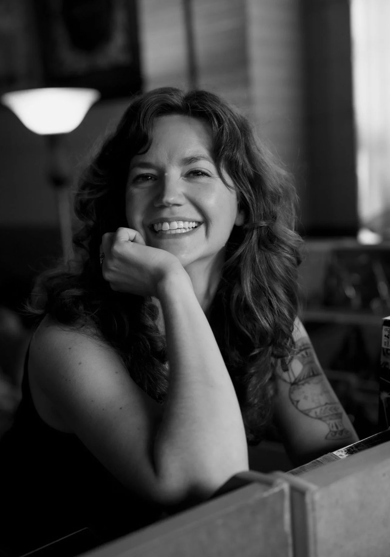 A smiling woman with curly hair resting her chin on her hand, sitting indoors with a tattoo sleeve on her left arm, black and white photo.