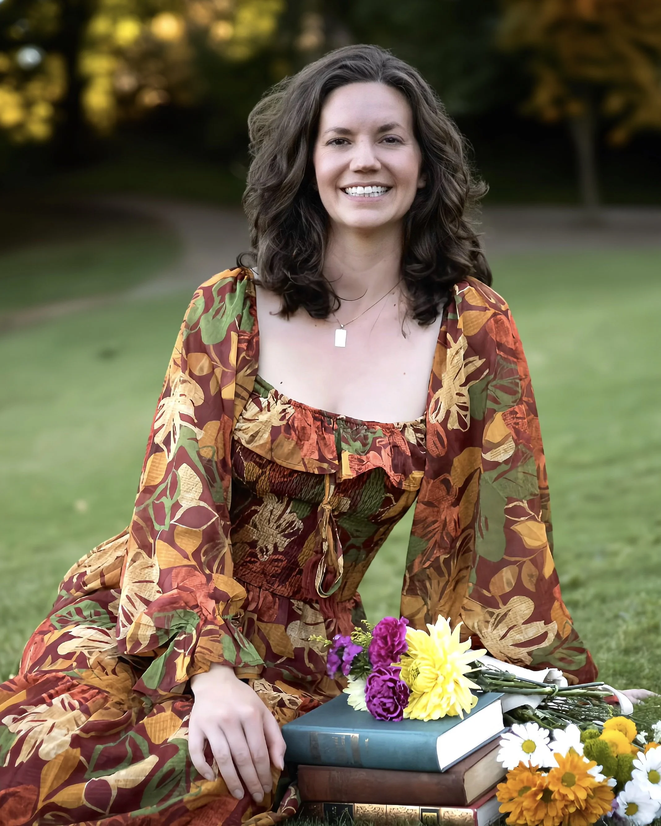 A woman with curly brown hair sits on the grass in a park, wearing a colorful floral dress. She is smiling and surrounded by flowers and books, with a blurred background of trees and greenery.
