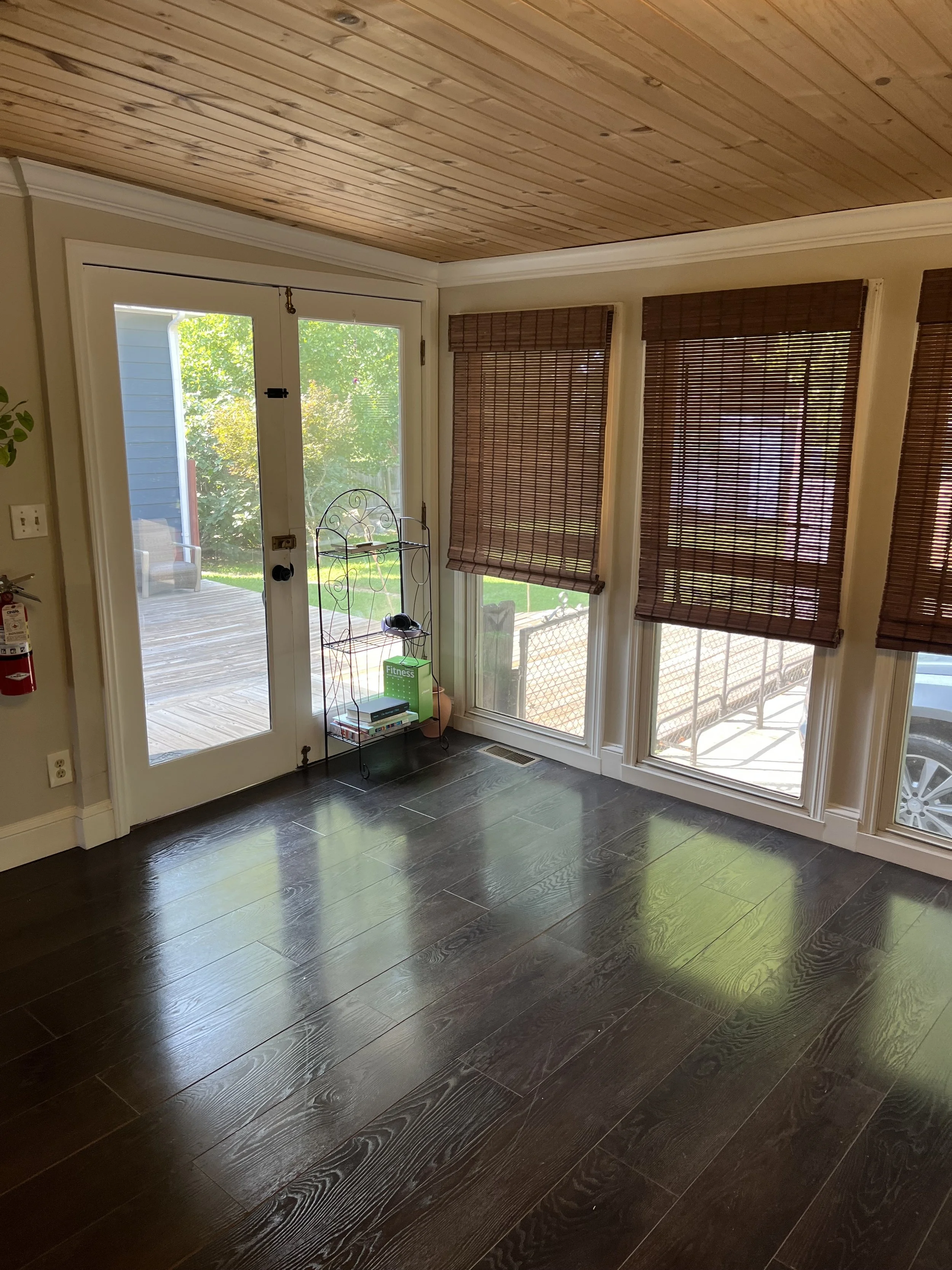 Interior view of a room with dark wood flooring, a glass door leading outside, and three windows covered with brown blinds.