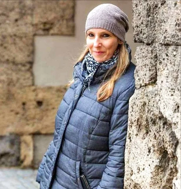 Eine Frau trägt eine graue Mütze, einen blauen gepufften Mantel und eine schwarze, weiße Schal und steht vor einer alten Stemwall. Sie lächelt leicht.