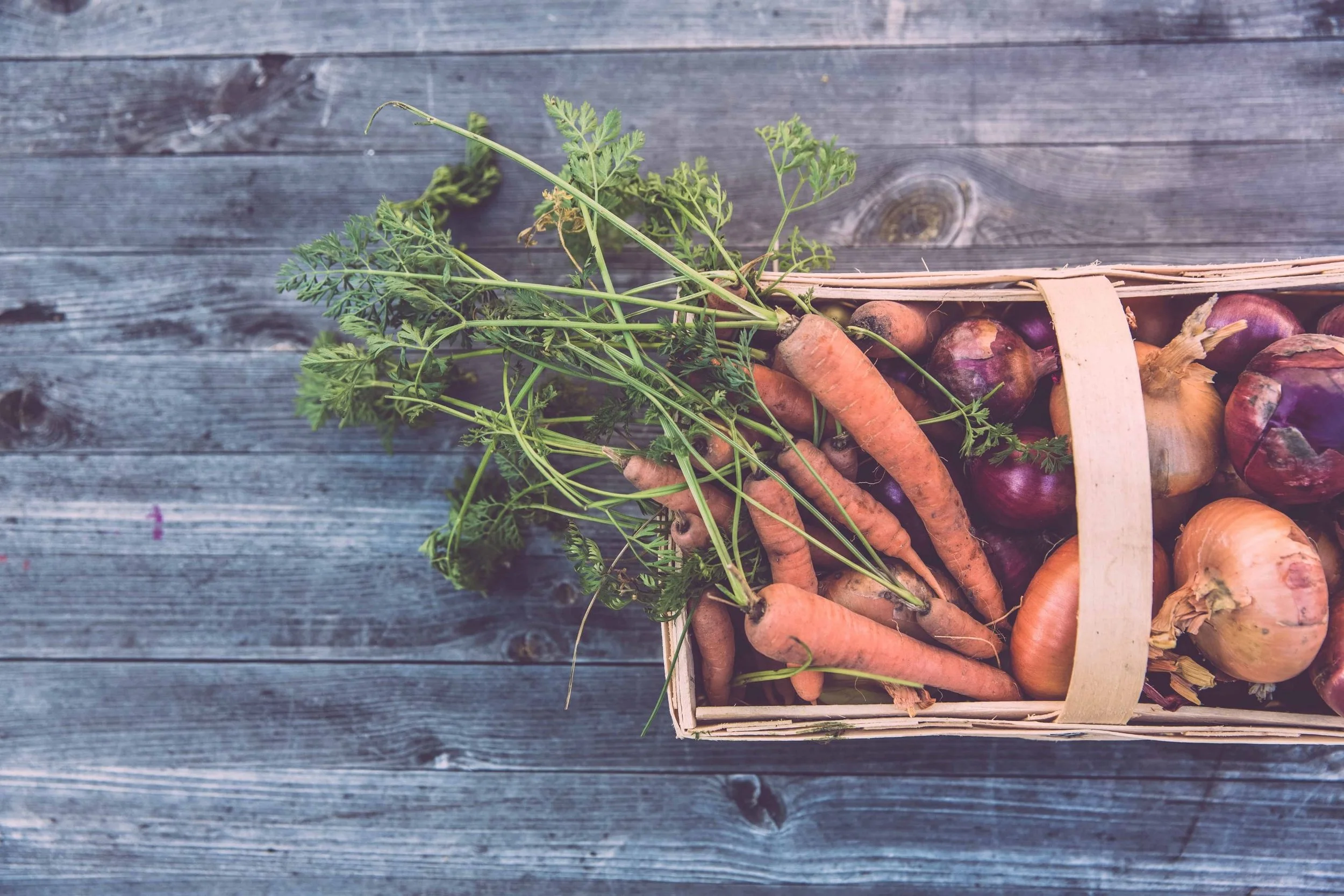 A wooden basket containing freshly harvested carrots and onions, placed on a weathered wooden surface.