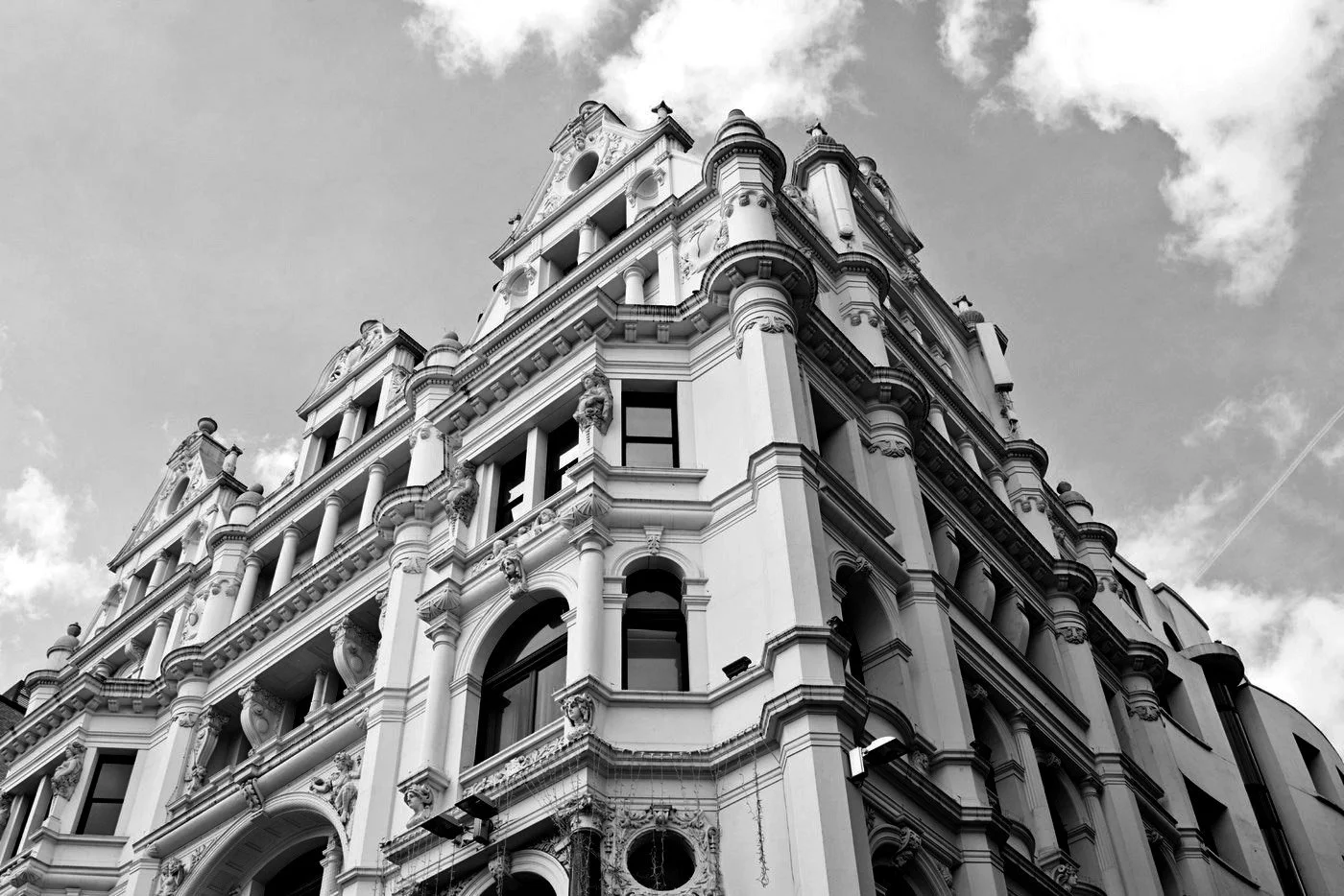 Black and white photo of an ornate historic building with intricate architectural details and multiple windows, set against a partly cloudy sky.
