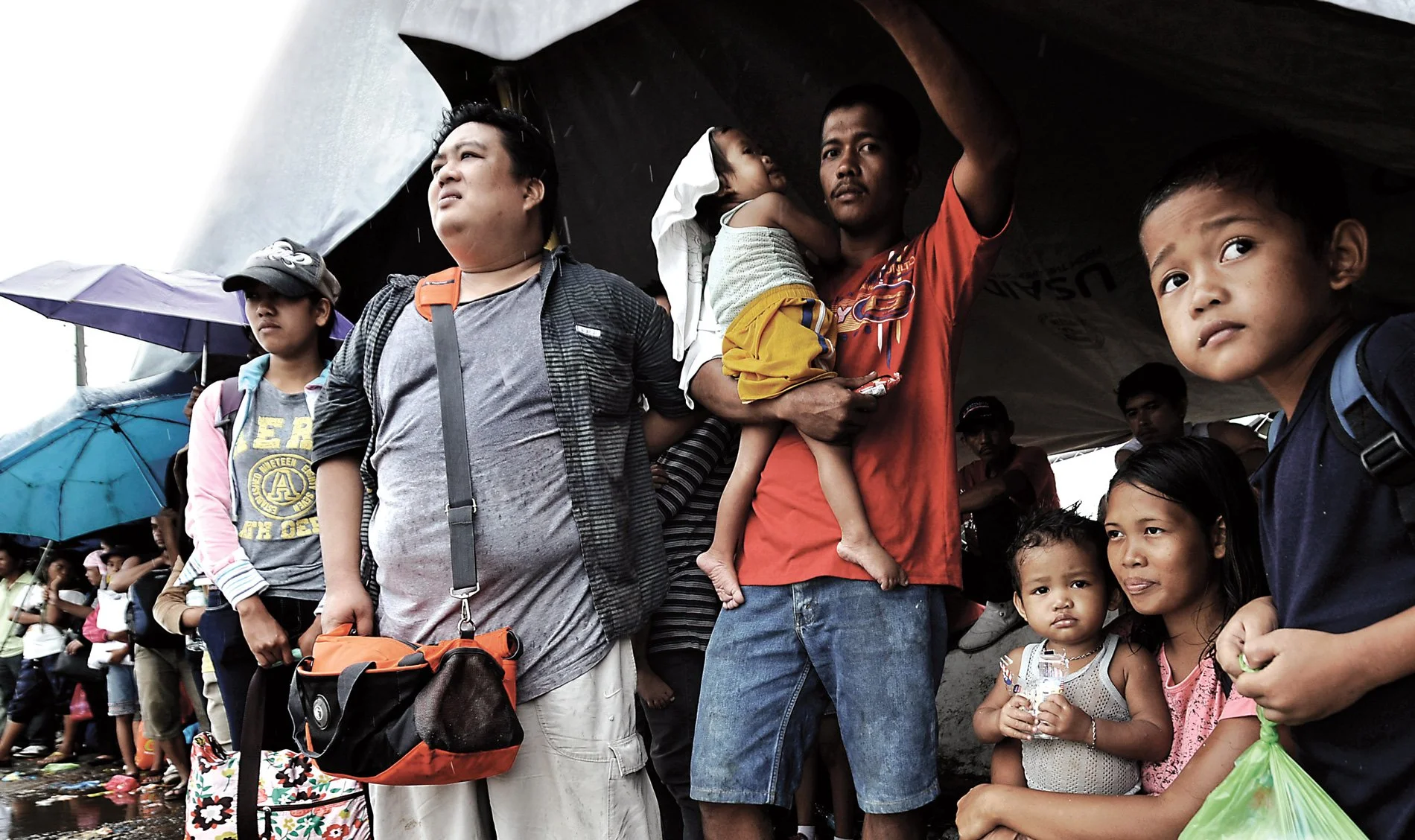 A group of refugees waiting under a shelter with umbrellas in a crowded area, including children and adults of diverse backgrounds.