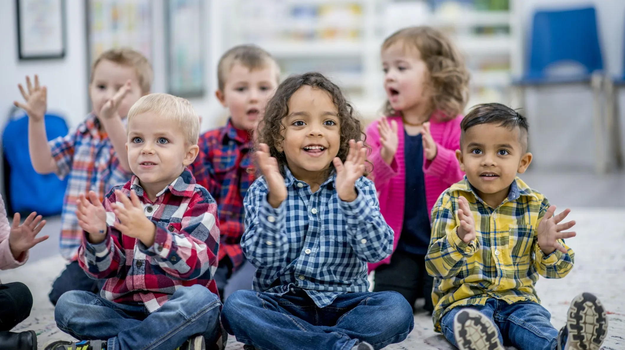 Group of young children sitting on the floor, clapping and smiling, in a classroom setting.