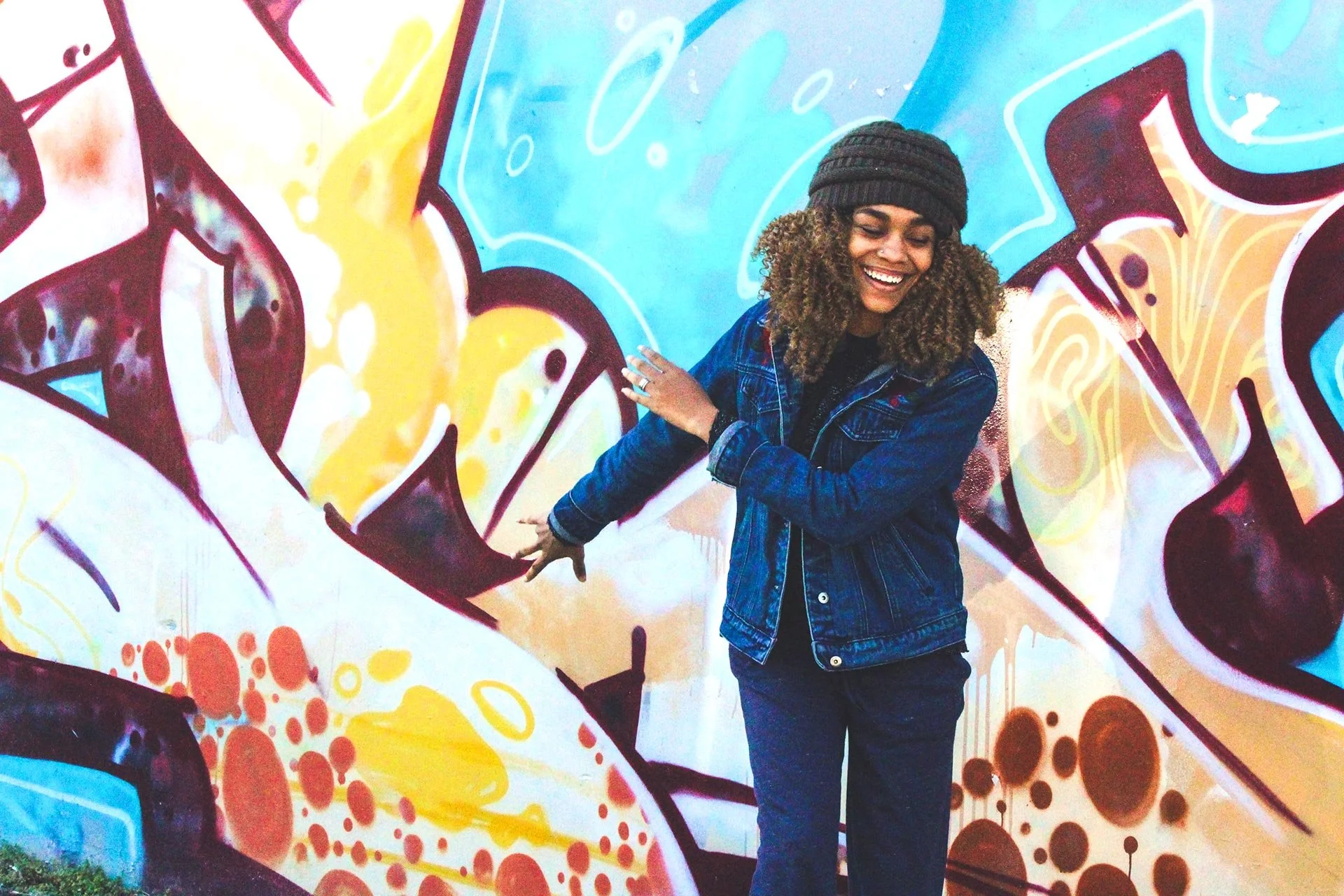 A woman with curly hair in a dark beanie and denim jacket smiling while standing in front of a colorful graffiti wall.