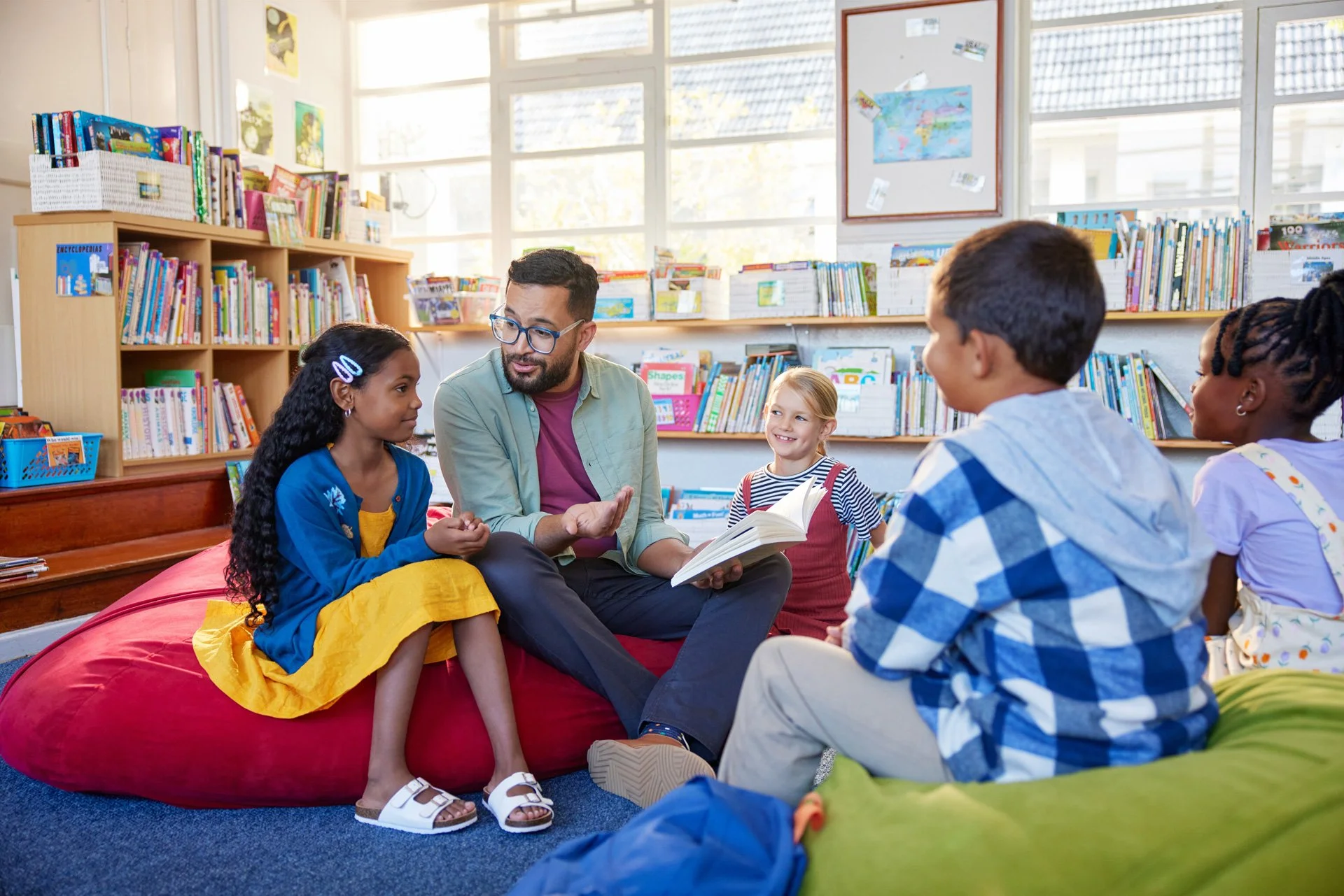 A man reading a book to a group of children in a library, sitting on the floor and a bean bag, with shelves of books and a window in the background.