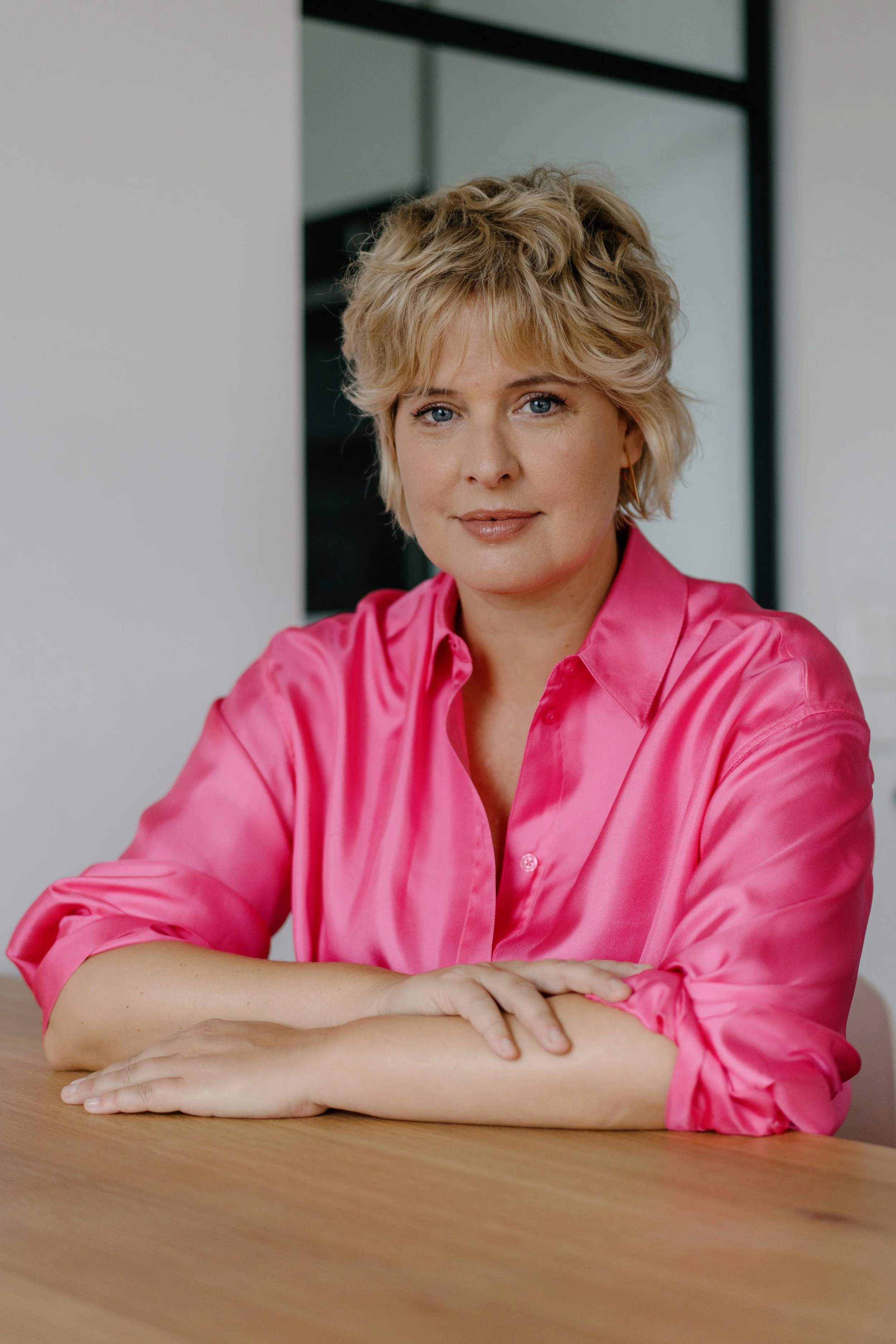 Tania Llasera in a pink blouse with arms on a table