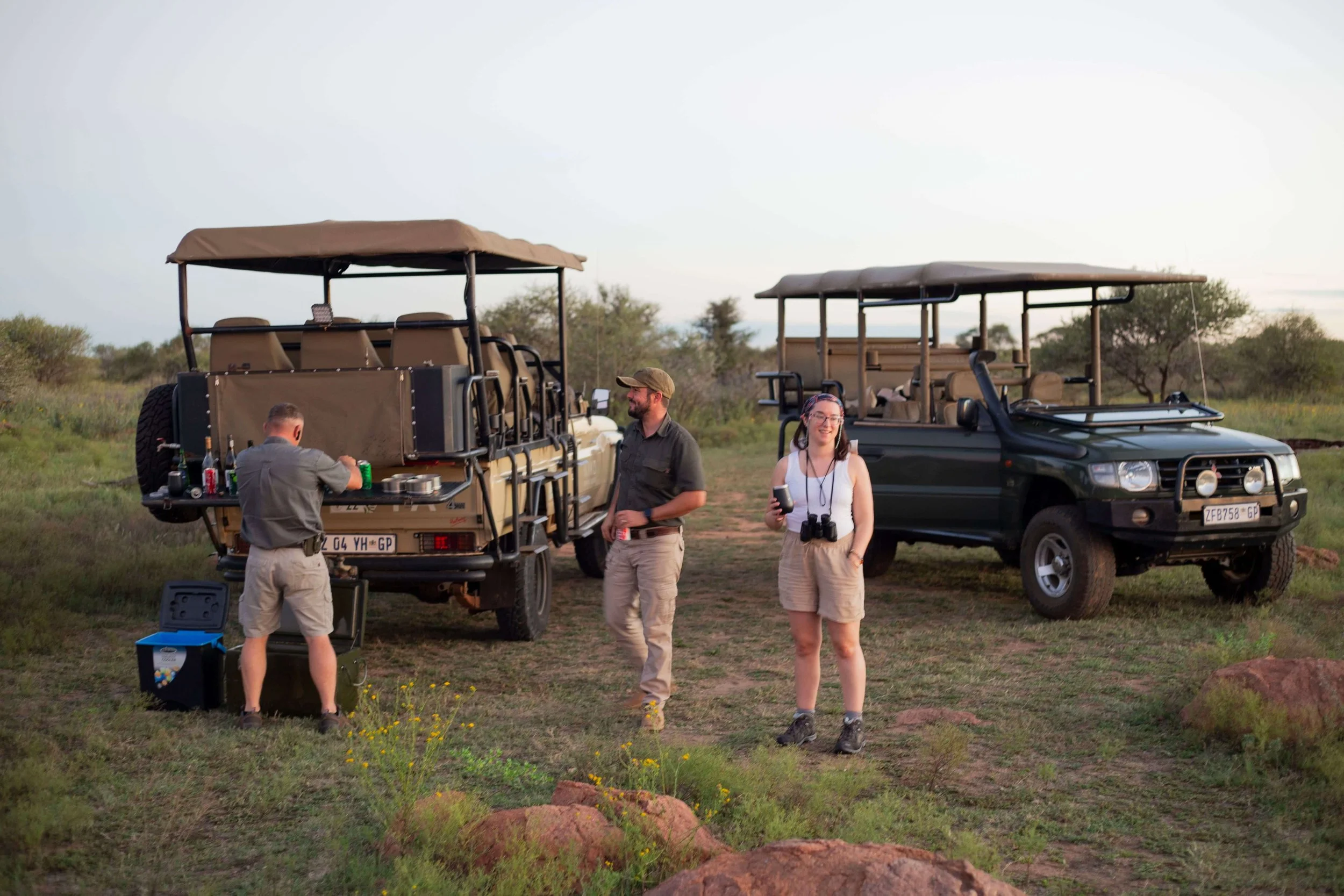 People preparing for a safari with open-top vehicles in a grassy savanna at sunset.