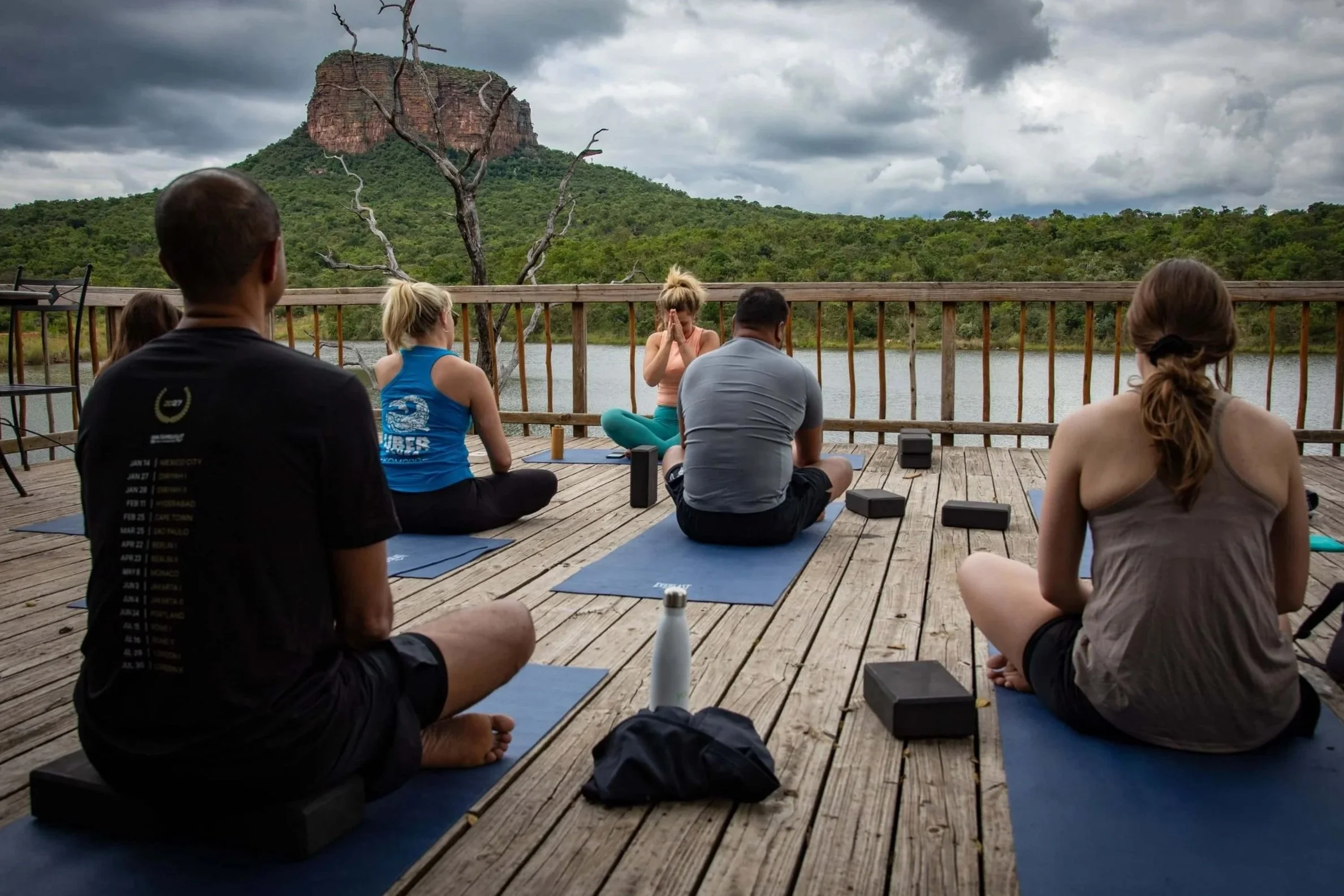 Group of people practicing yoga outdoors on a wooden deck with a mountain landscape and cloudy sky in the background.