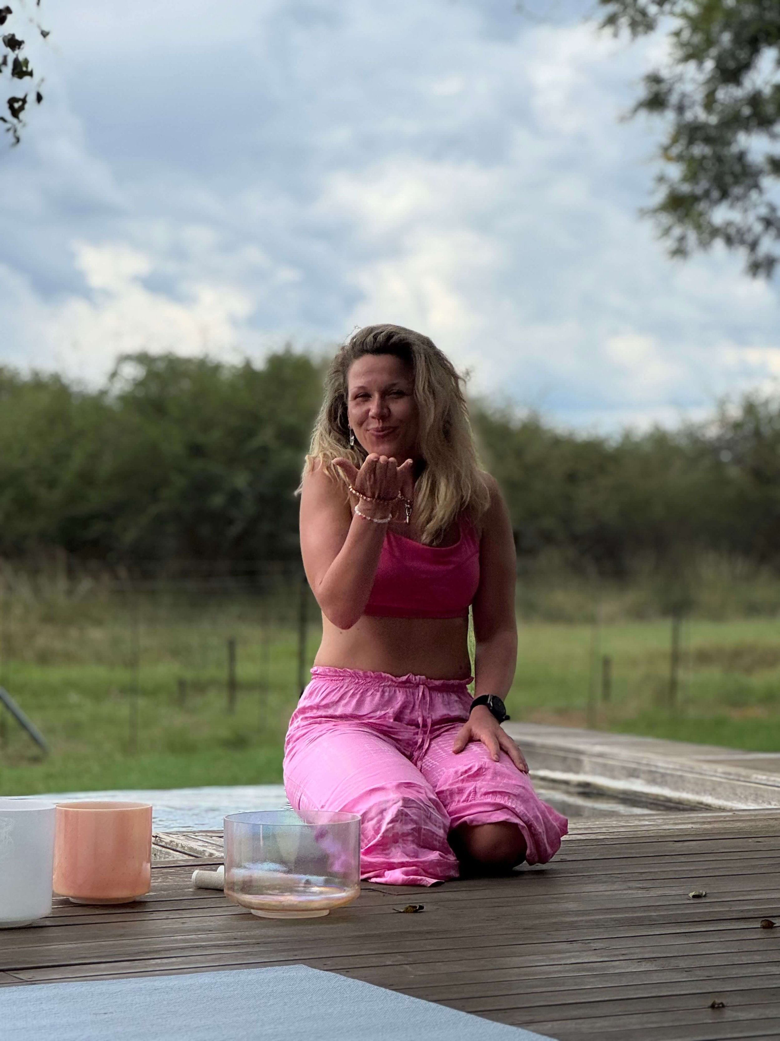 A woman with wavy blonde hair blowing a kiss towards the camera while seated outdoors on a wooden deck, with a grassy background and cloudy sky.