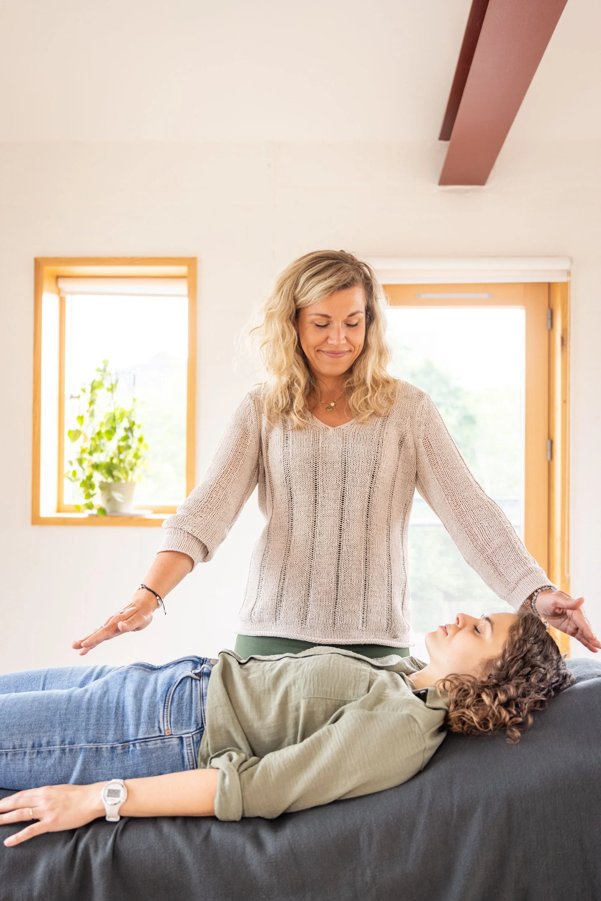 A woman with curly hair lying on a massage table with her eyes closed, while a female therapist in a beige sweater performs a healing or relaxation technique in a bright room with windows.