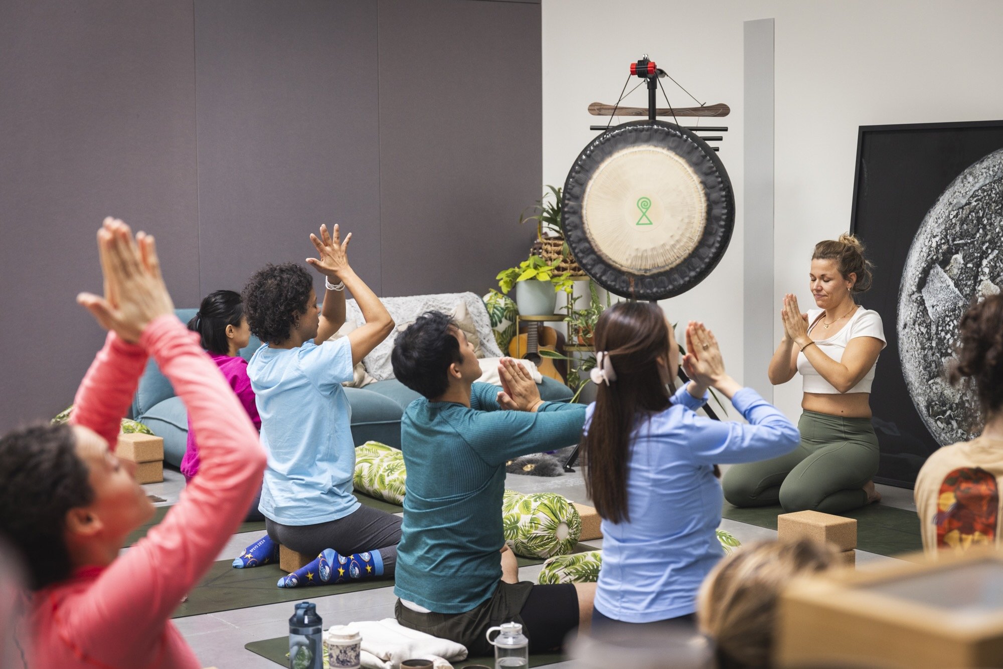 A group of people participating in a yoga class, sitting on yoga mats with some raising their hands, in a room decorated with plants and artwork, with an instructor leading the session.