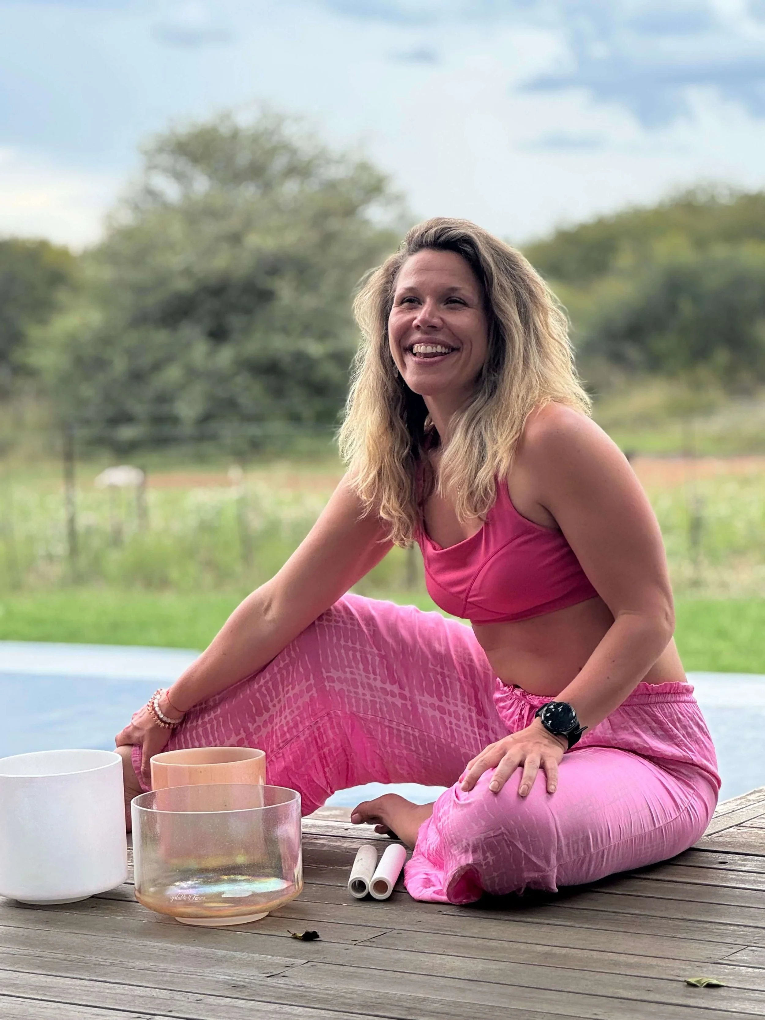 A woman in pink yoga attire sits on a wooden deck outdoors, surrounded by outdoor singing bowls and white and clear glasses, with a blurred natural background of trees and a cloudy sky.