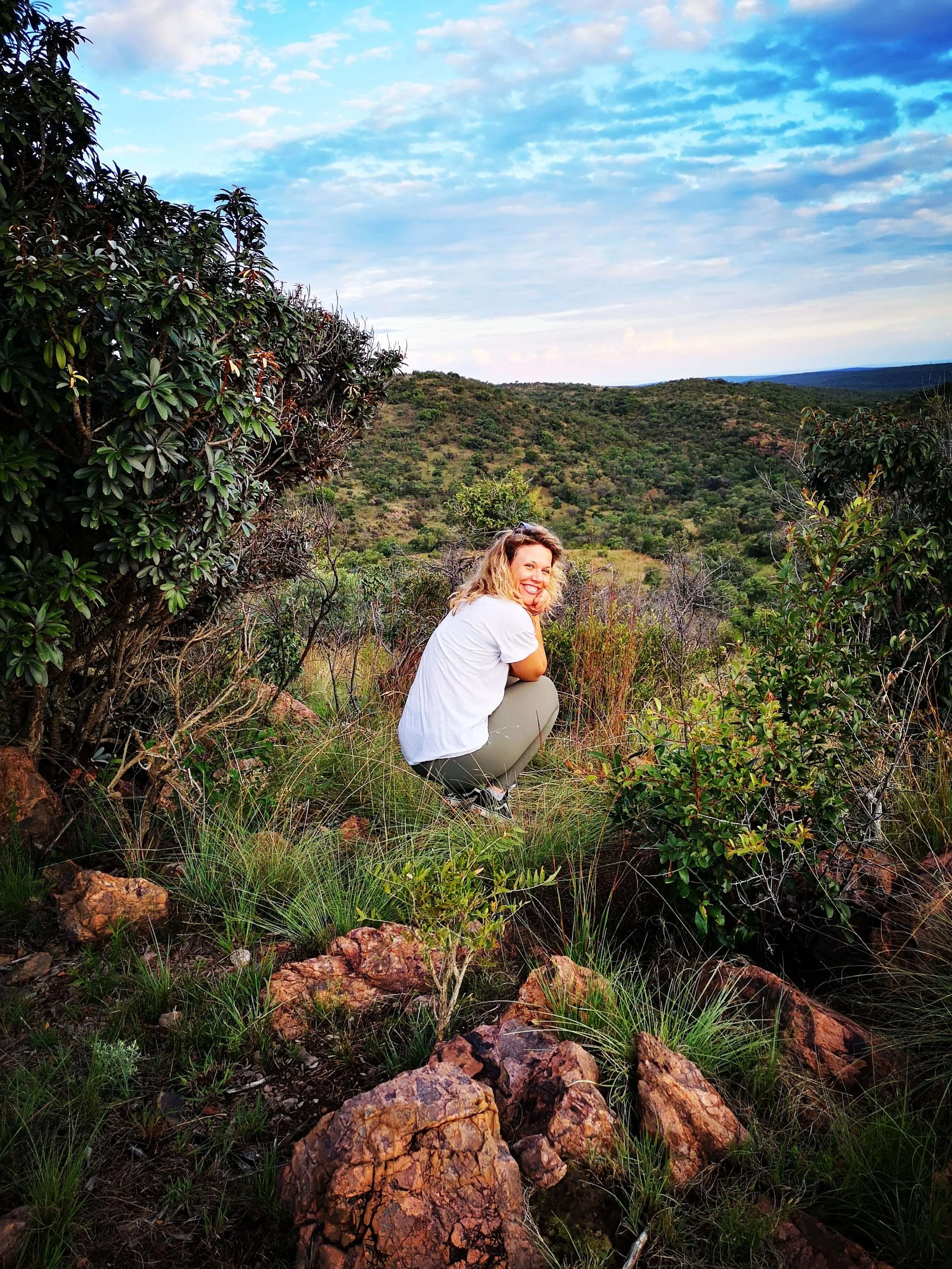 A smiling woman with curly blonde hair, wearing a white shirt and tan pants, crouches among rocks and green bushes on a hilltop with a view of rolling hills covered with trees under a partly cloudy sky.