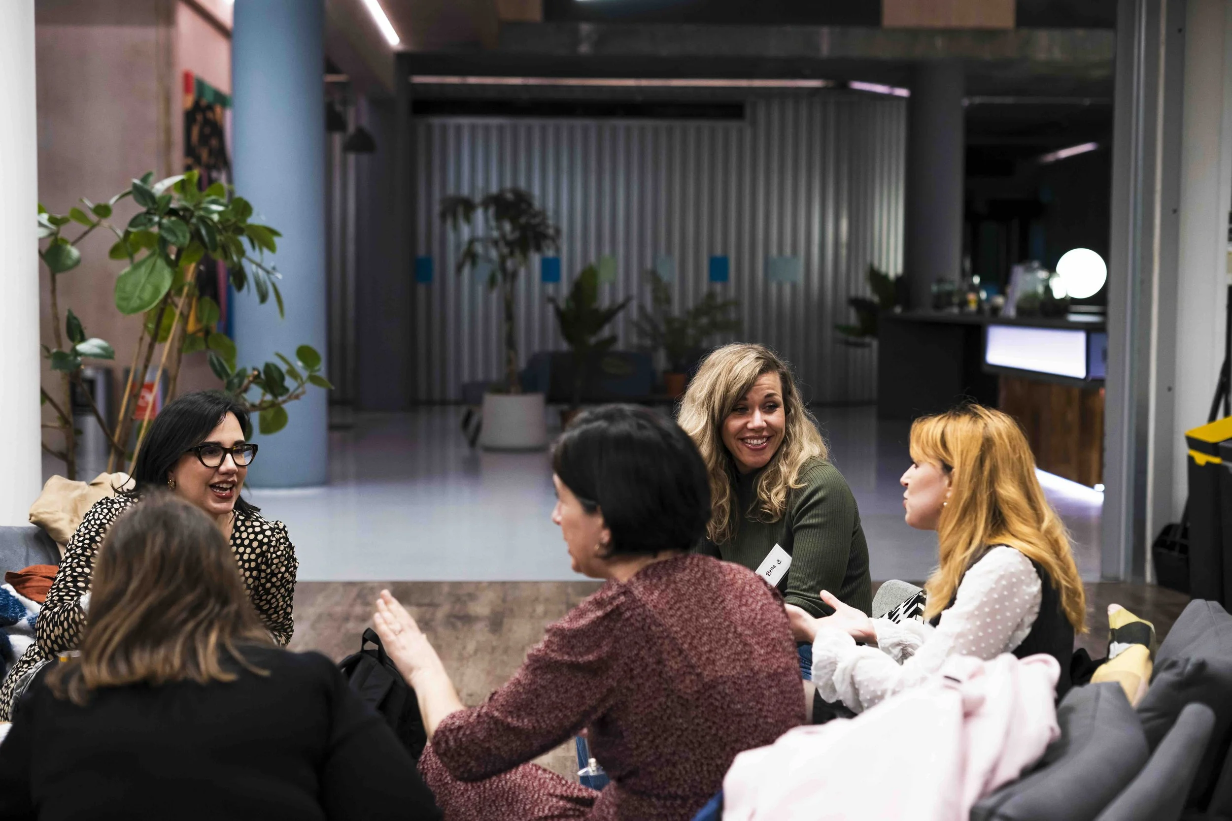 A group of diverse women sitting in a circle engaged in conversation at an indoor gathering, with some smiling and gesturing.