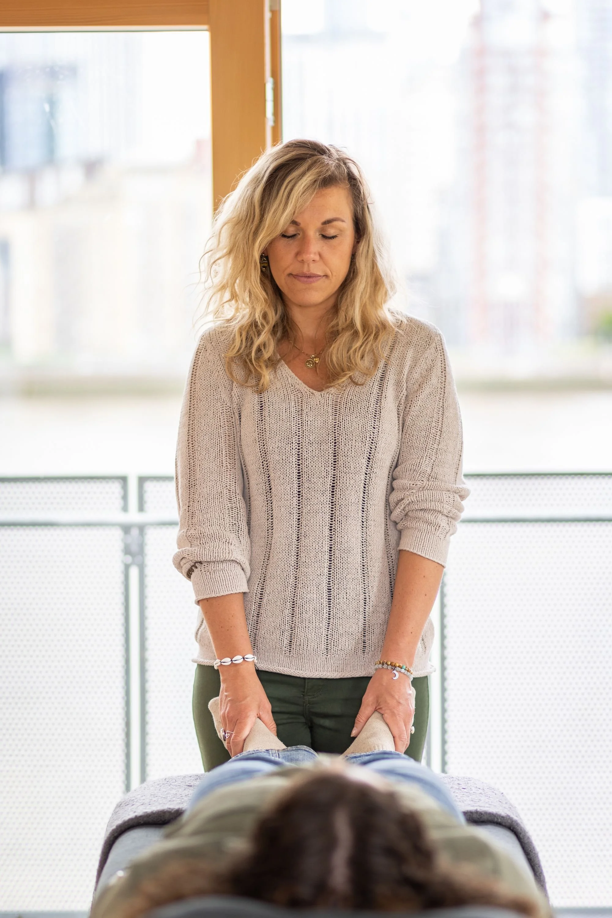 A woman with blond, curly hair standing in front of a person lying face down, holding their ankles. The woman is wearing a beige knit sweater and various bracelets, with a neutral expression, in a brightly lit indoor setting with windows in the background.