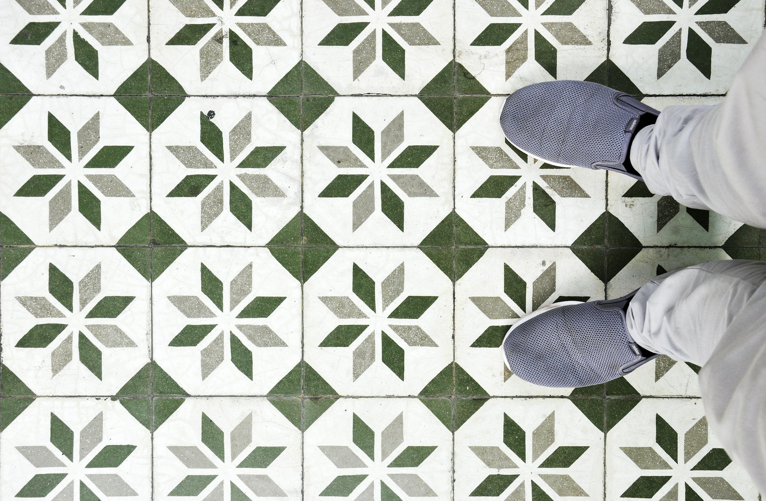 Looks down at a tiled floor featuring geometric green and white patterns, with a person's feet in gray sneakers and beige pants visible at the edges.