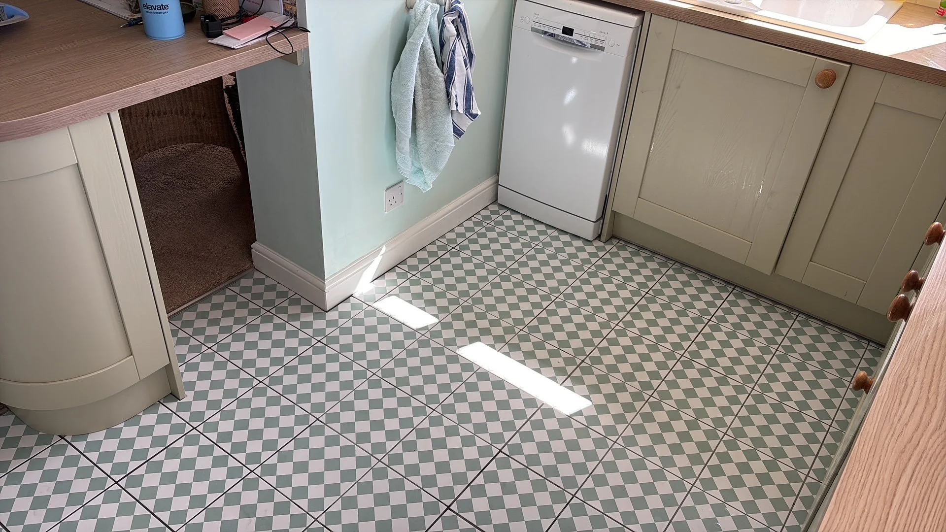 A kitchen corner featuring light green cabinets with wooden knobs, a dishwasher, a patterned tiled floor, a towel hanging on the wall, a countertop with various items, and sunlight reflecting on the floor.