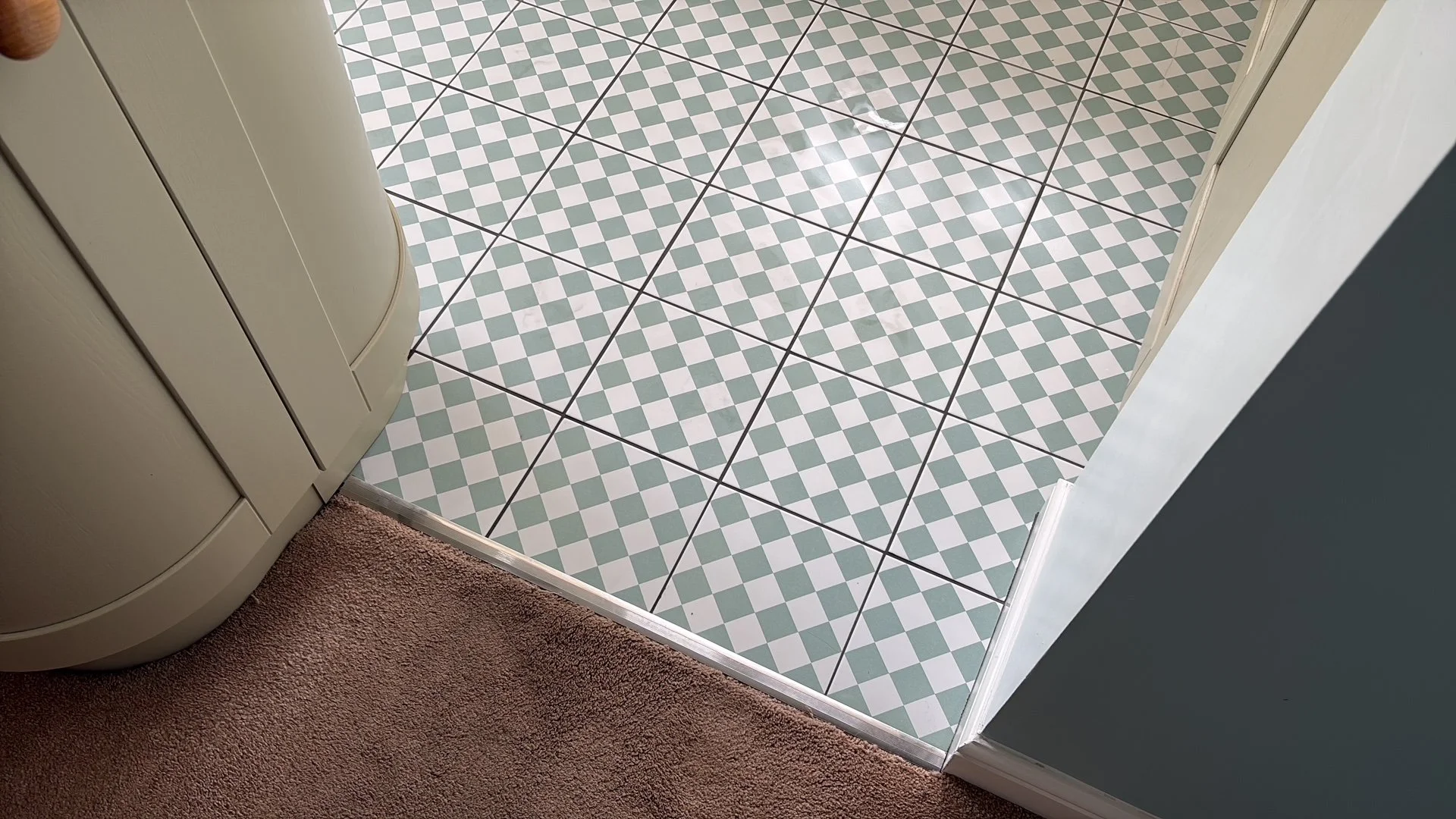 Bathroom floor with green and white diamond pattern tiles, transitioning to carpeted area, with part of a white curved cabinet or appliance on the left and a door frame on the right.