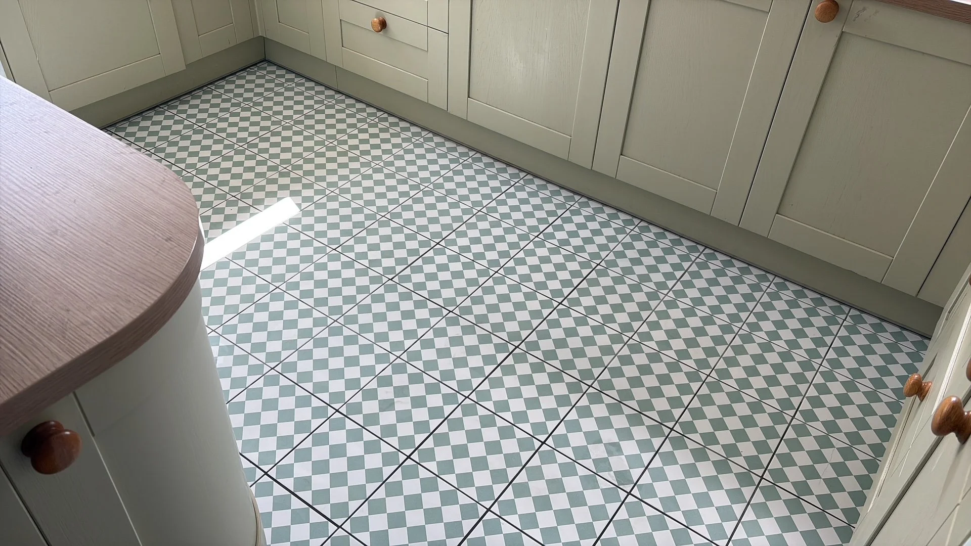 Kitchen floor with green and white checkered tiles, cream-colored cabinets with wooden knobs, and a wooden countertop edge visible on the left.