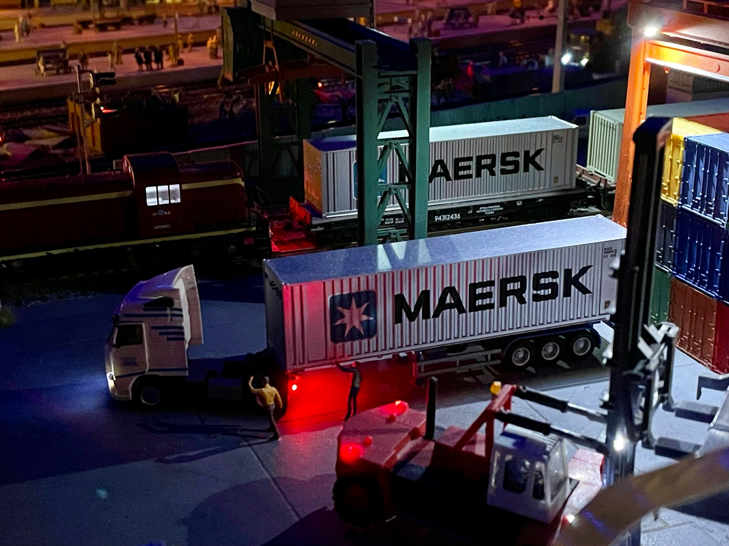 A nighttime scene of a freight train yard with stacked shipping containers, some labeled Maersk, and a few small figures of people near the trains.