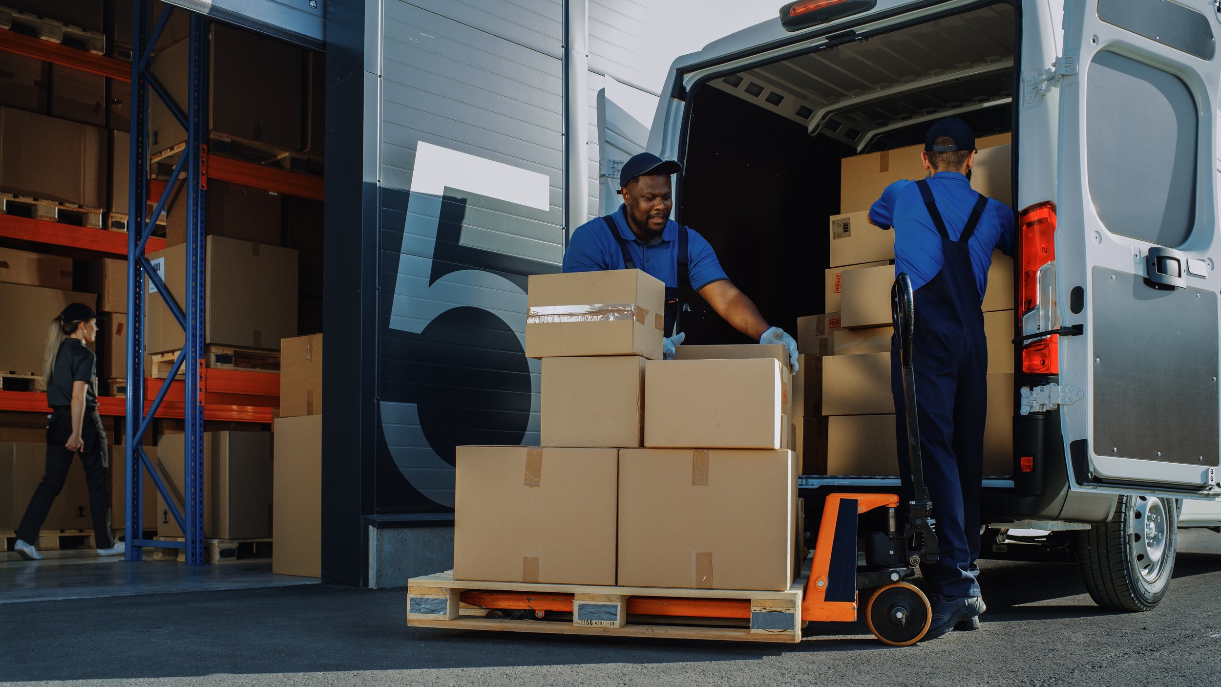 Two workers loading boxes into a white delivery truck in a warehouse parking lot, with a woman walking nearby and shelves filled with boxes in the background.