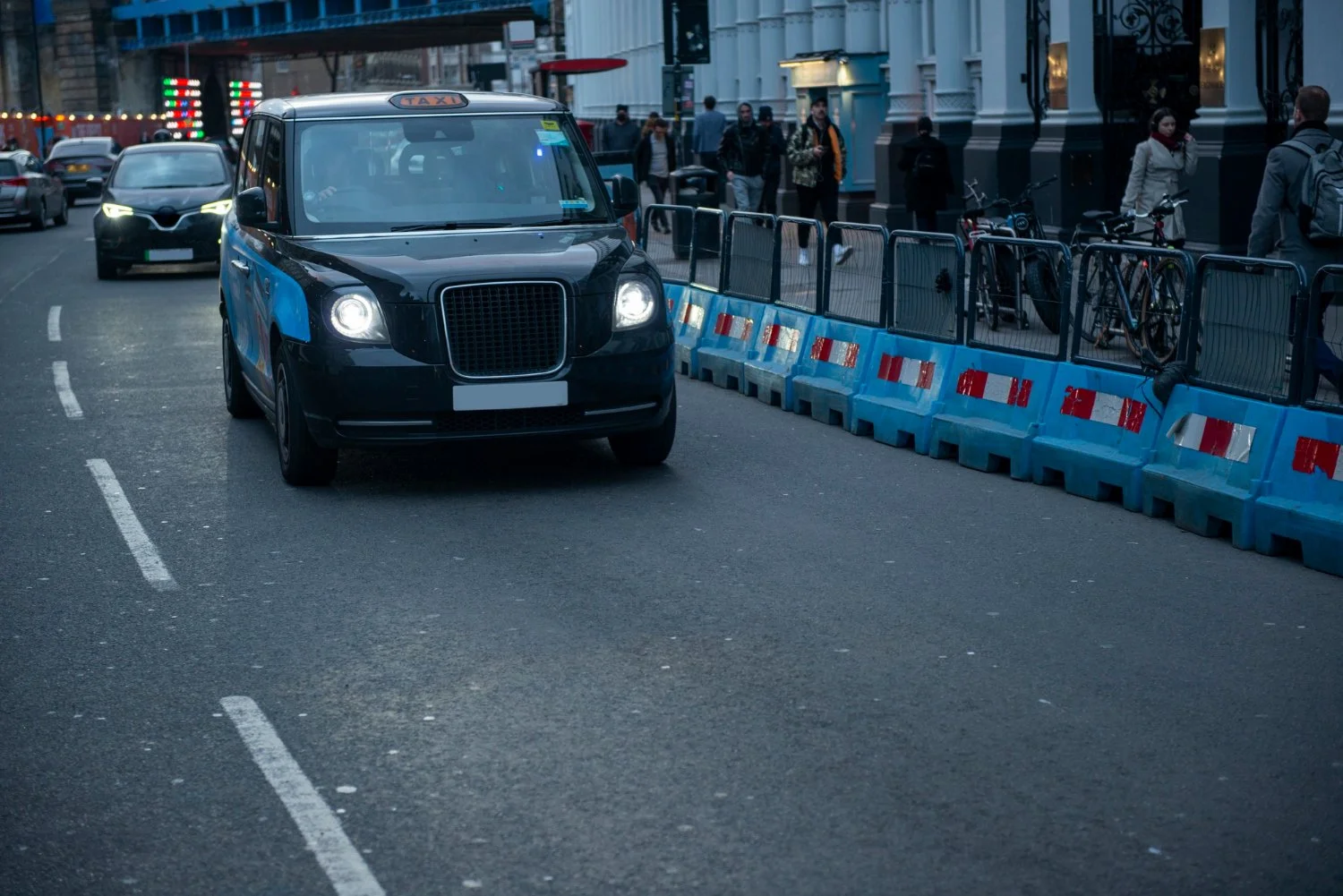 A black taxi cab on a city street near a sidewalk with pedestrians and bicycles separated by blue and orange street barriers.