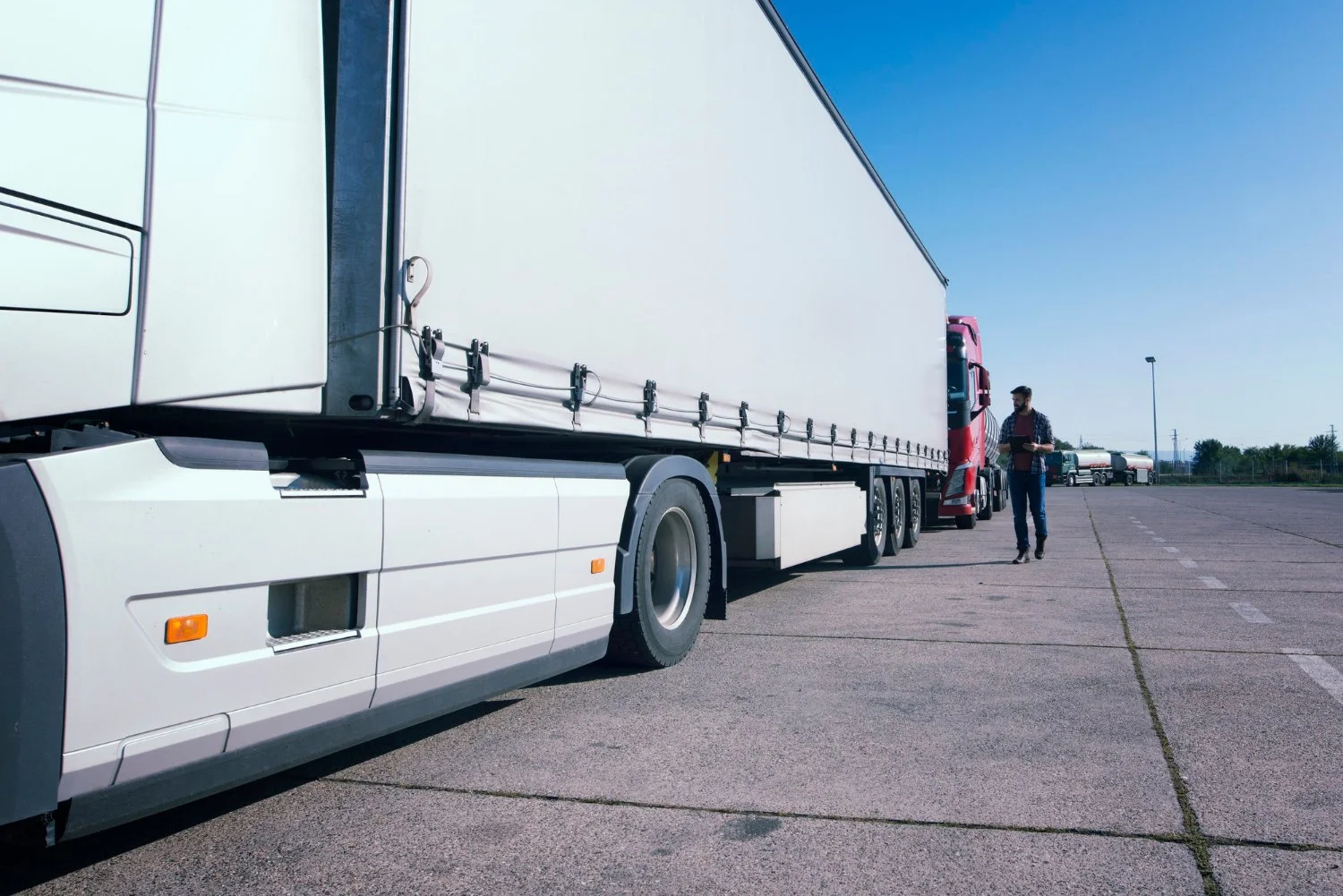 A man walking near a large white semi-truck parked in an empty lot under a clear blue sky.