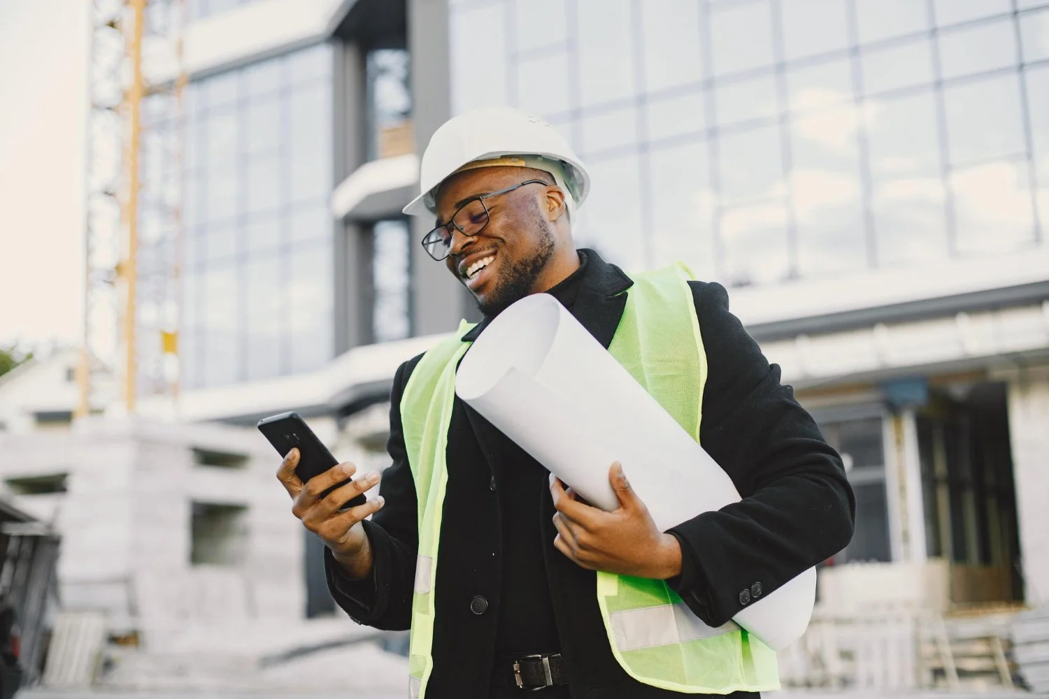 A smiling man in a hard hat and safety vest holding rolled blueprints and looking at his phone at a construction site with a modern glass building in the background.