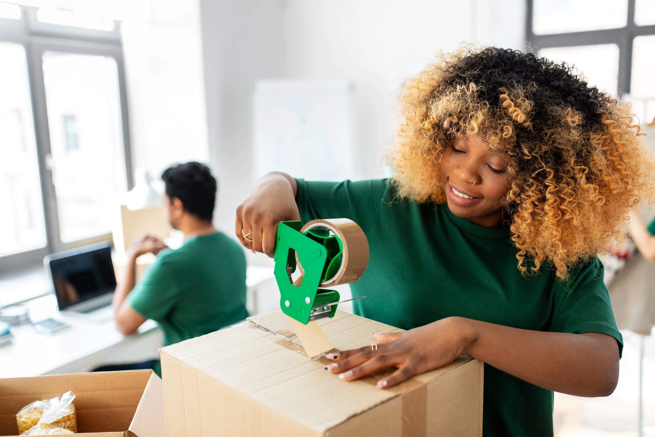 A woman with curly black and blonde hair using a tape dispenser on a cardboard box in an office setting.
