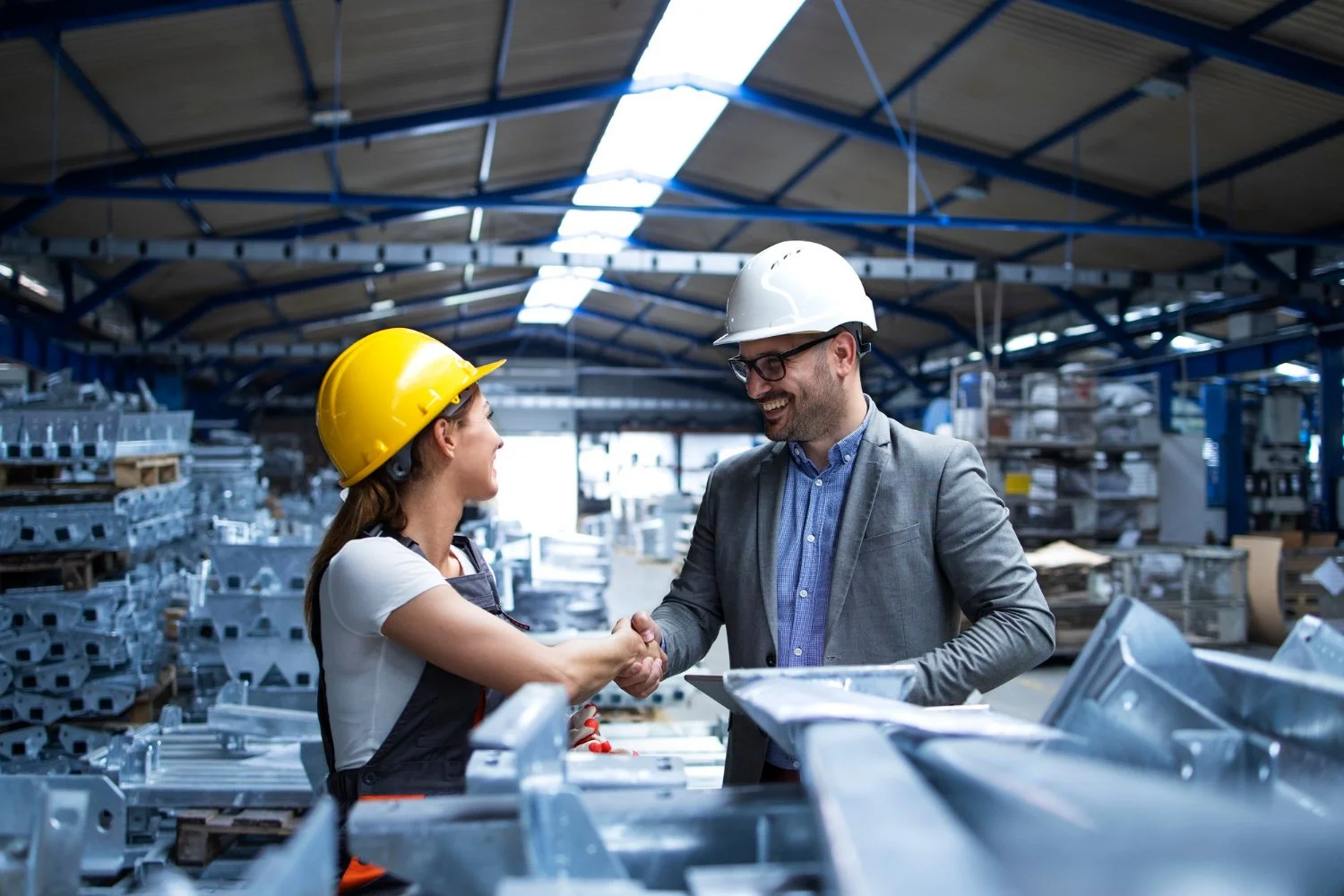 Two workers wearing yellow and white safety helmets shaking hands in an industrial warehouse surrounded by metal parts.