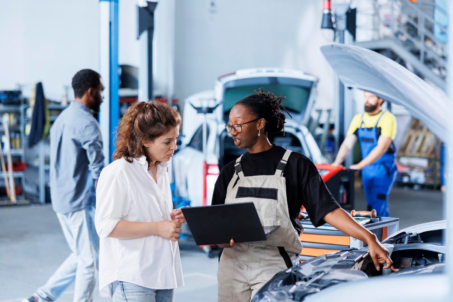 Auto mechanic explaining car engine to woman customer in auto repair shop, with another mechanic working in the background.