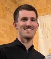 A man with short dark hair and a beard, smiling, wearing a black shirt, standing in front of a yellow wall with writing.
