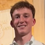 A smiling young man with short curly hair wearing a white shirt, standing in front of a yellow backdrop.