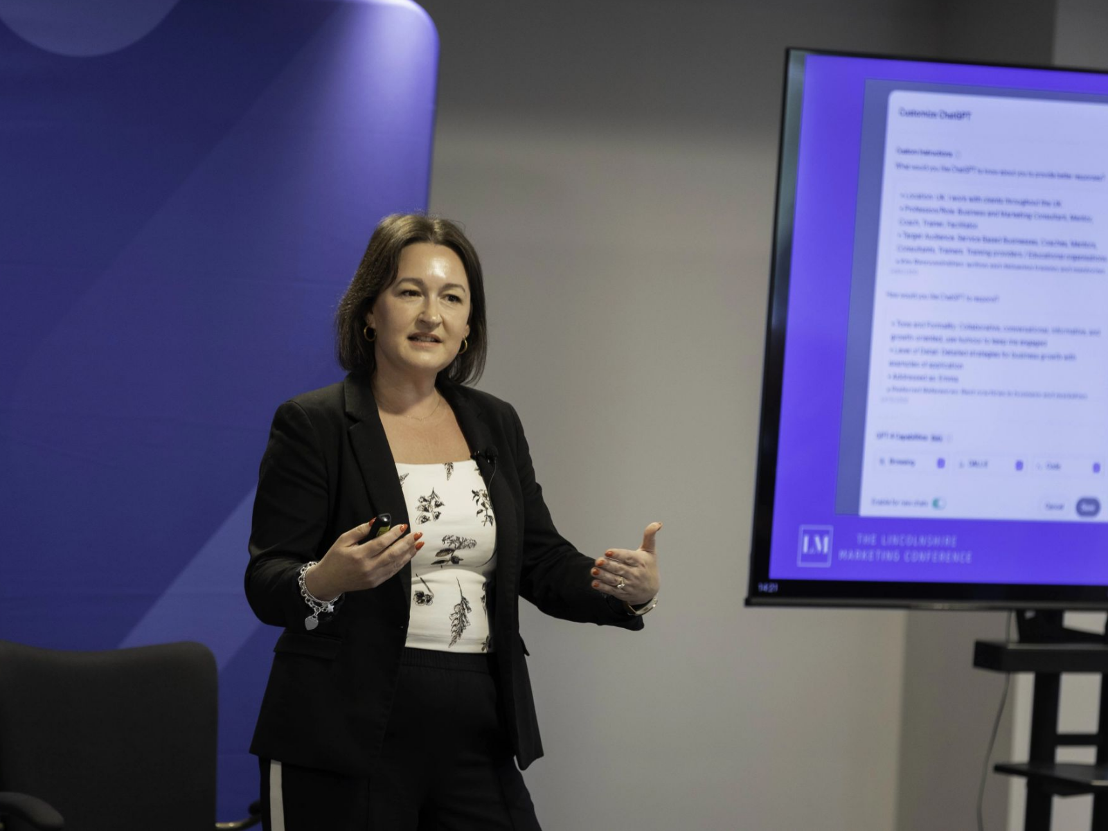 Emma Ellse, founder of AI Business Growth Academy, is giving a presentation on AI Adoption to business leaders in front of a blue wall and a large monitor displaying a document.